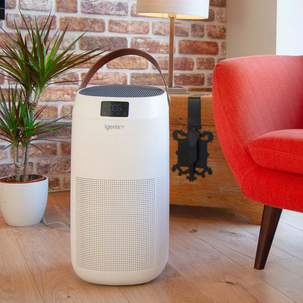 A white Igenix air purifier with a digital display sits on a wooden floor next to a green potted plant, orange armchair, and a wooden trunk in a cozy living room with brick walls.
