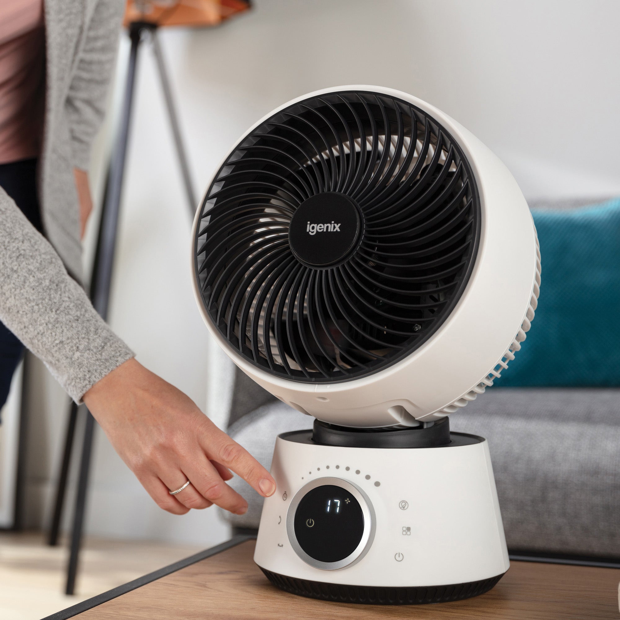 A person’s hand is pressing a button on a white Igenix tabletop fan with a digital display, placed on a wooden surface in a modern living room.