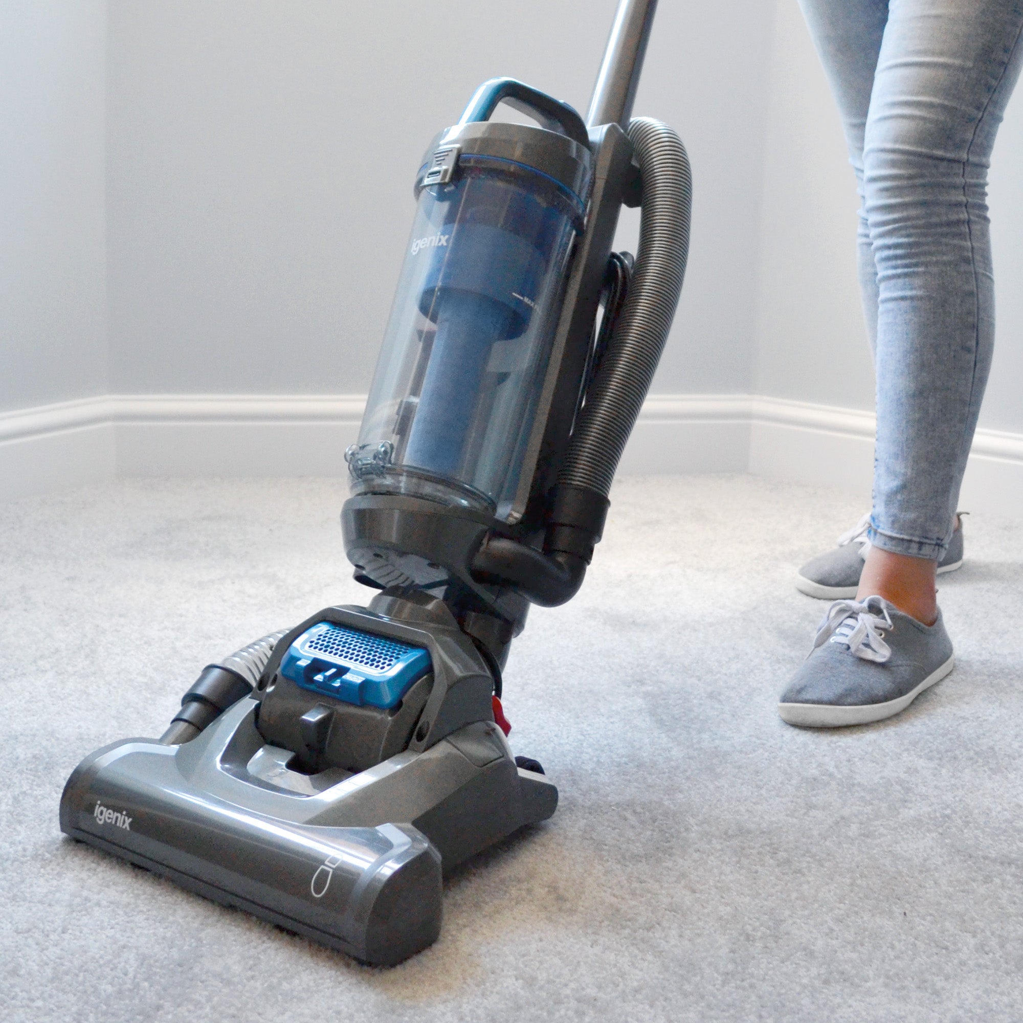 Person vacuuming a light gray carpet with a gray upright vacuum cleaner in a bright room. Only their legs in jeans and gray shoes are visible.