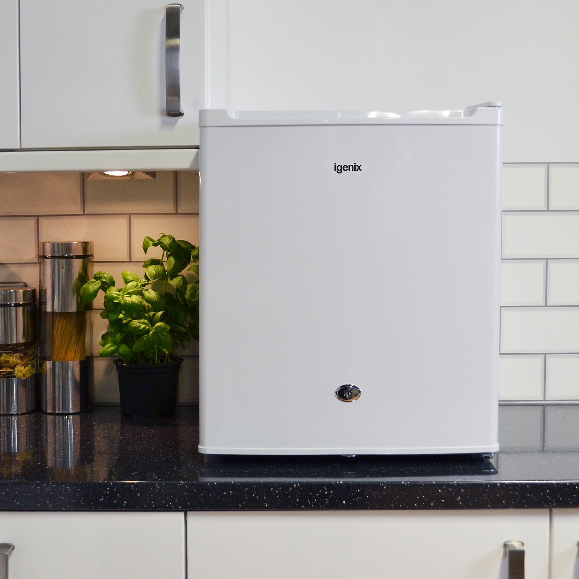 A white Igenix mini fridge sits on a black countertop in a modern kitchen, next to a plant and metal containers, with white cabinets and tiled backsplash in the background.