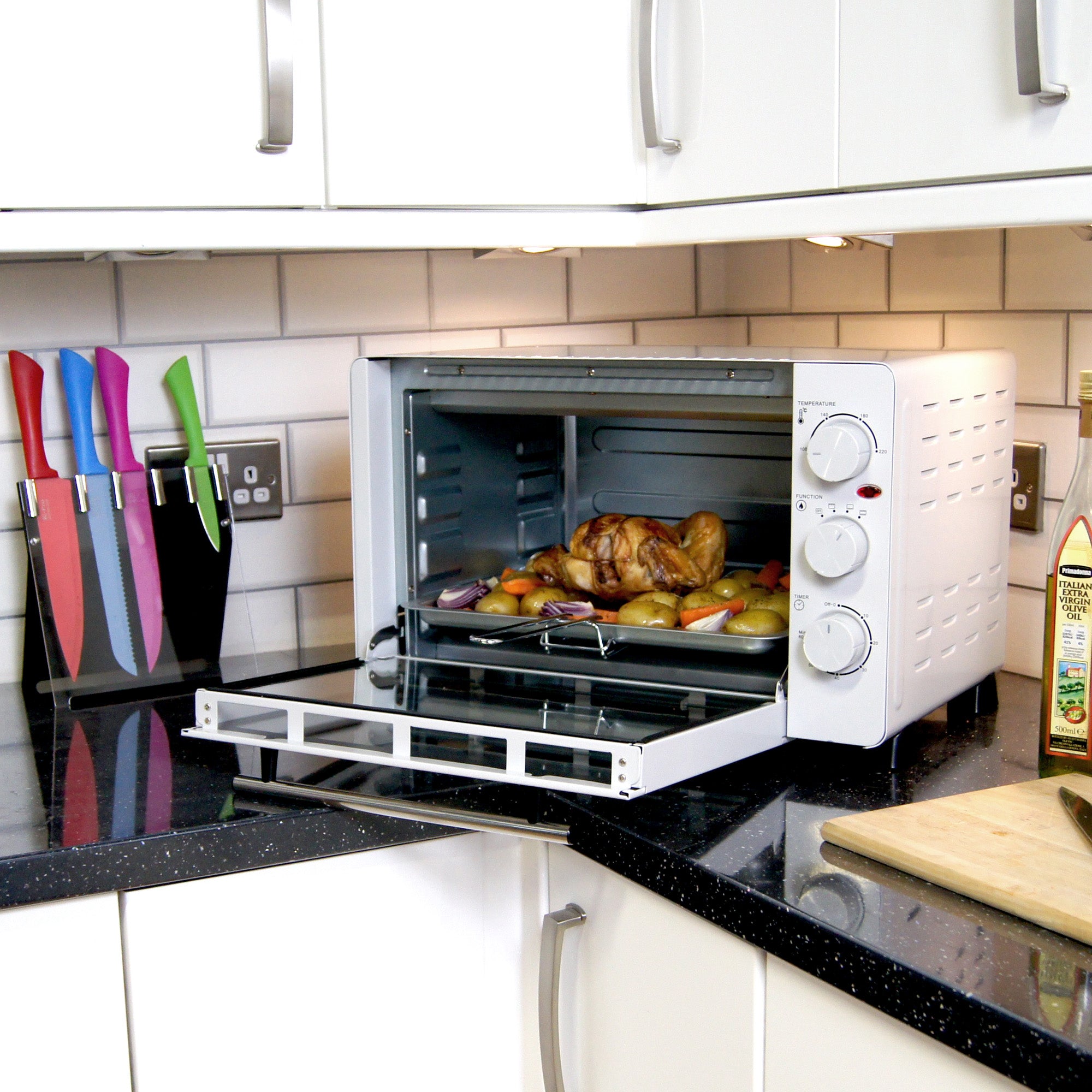 A countertop oven on a kitchen counter is roasting a chicken with vegetables. Colorful knives are in a holder nearby, and a wooden cutting board sits beside the oven.