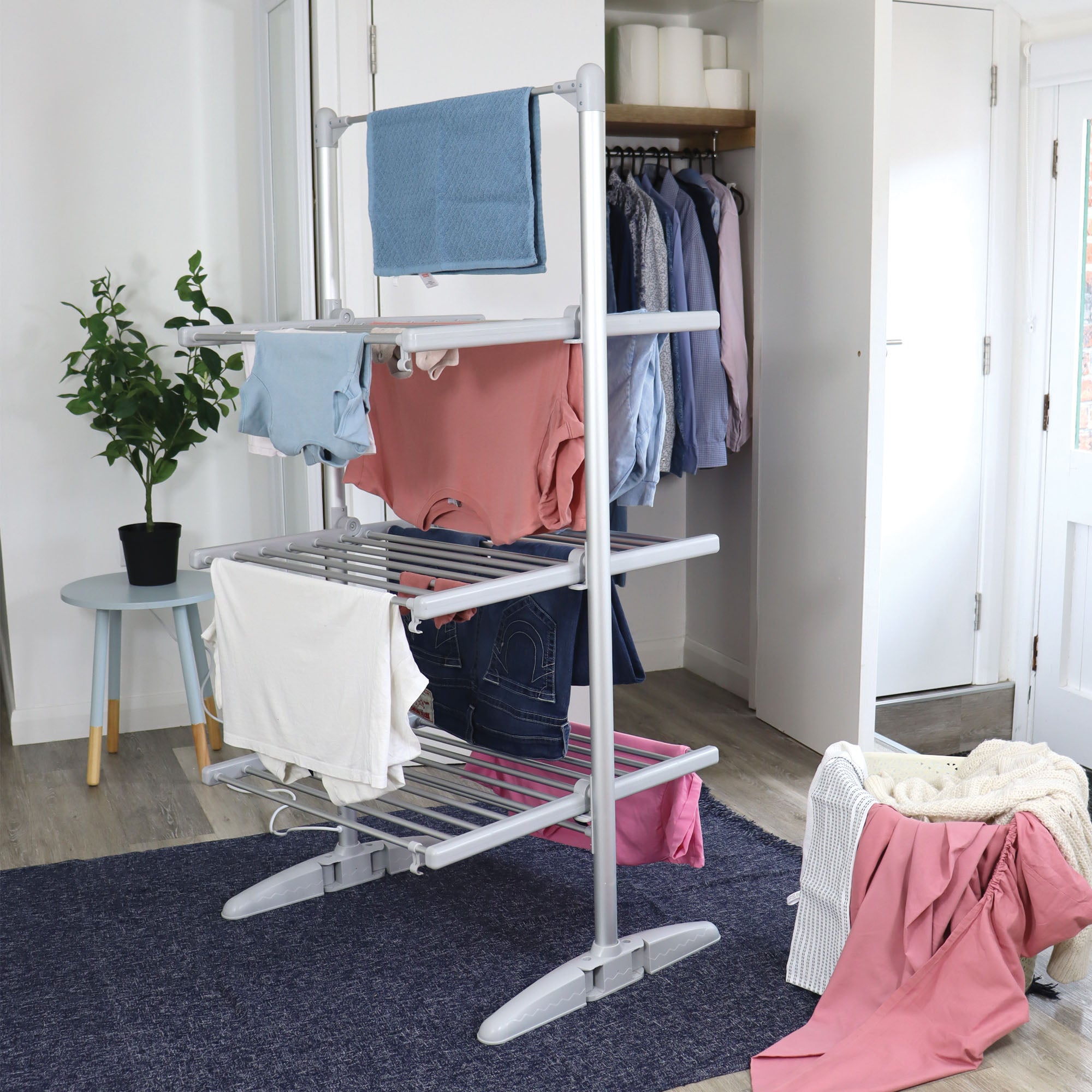 A laundry drying rack with clothes hanging on it stands in a tidy room with a blue rug, potted plant, side table, and an open closet. A pile of laundry sits on a chair nearby.