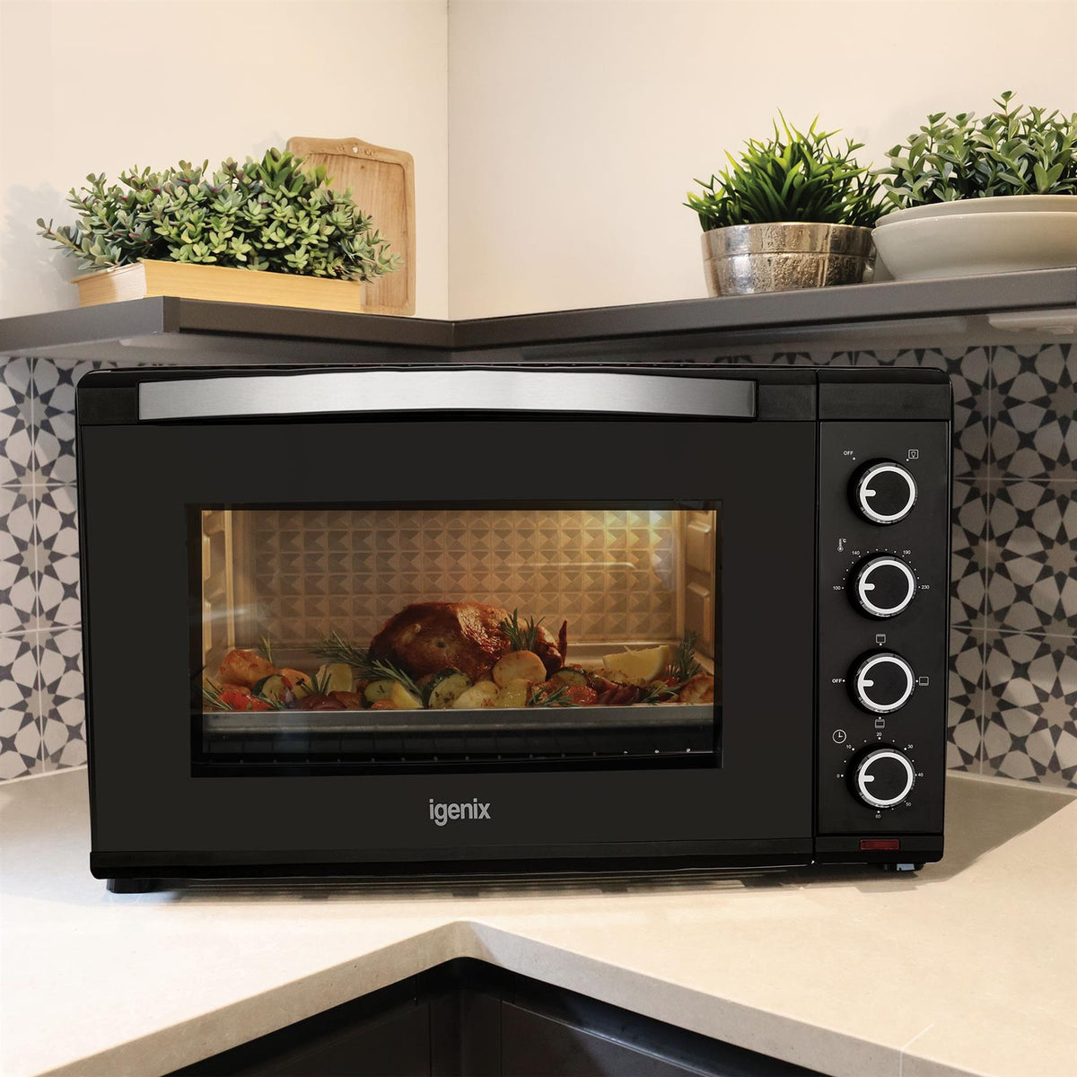 Black Igenix 60L Mini Oven with a glass door revealing a roasted chicken and vegetables inside, featuring four control knobs on the right side.