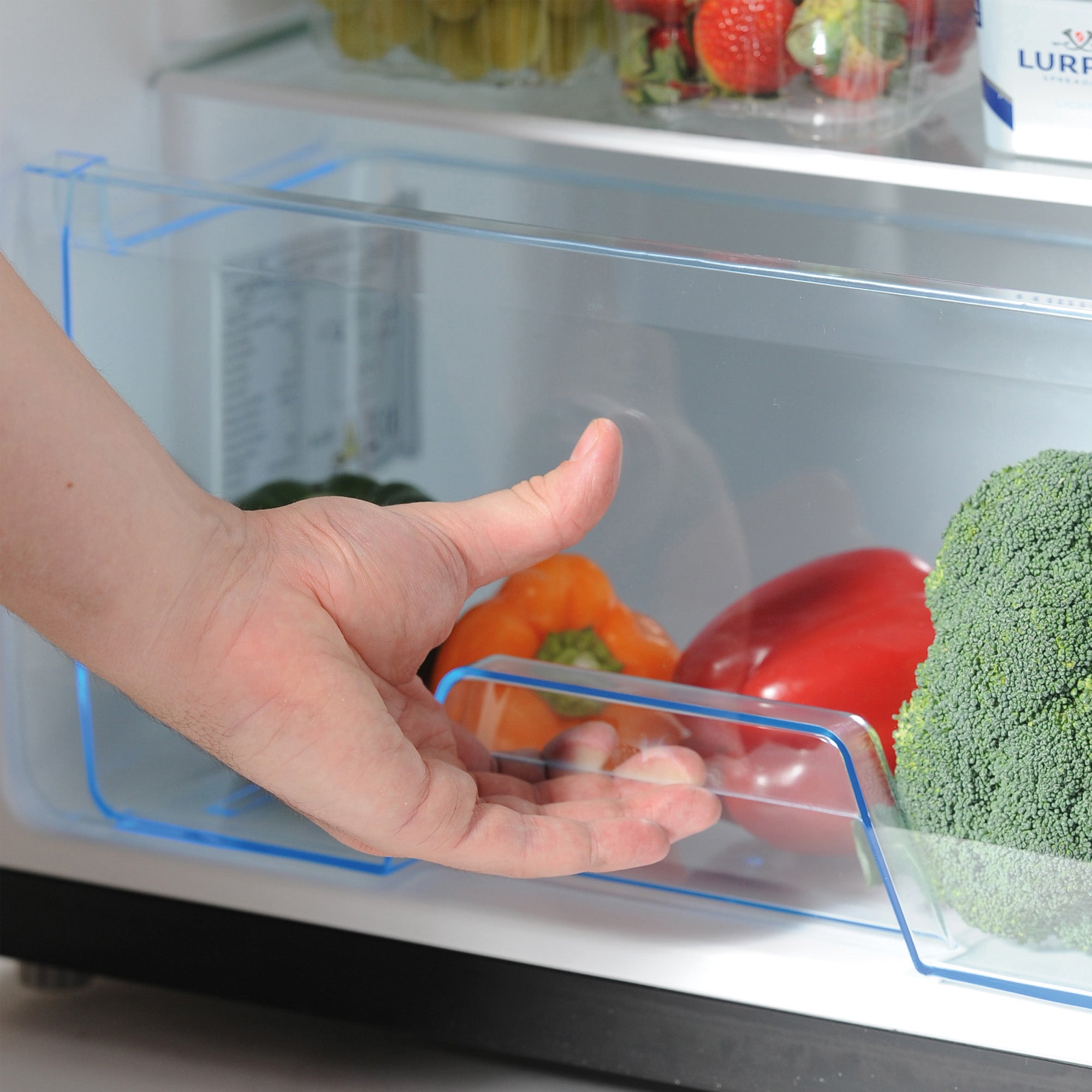 A hand reaches into the Igenix Under Counter Fridge with Ice Box (Black), accessing a compact drawer filled with broccoli, a red pepper, and an orange pepper, while other vegetables and food items are visible on the shelf above.