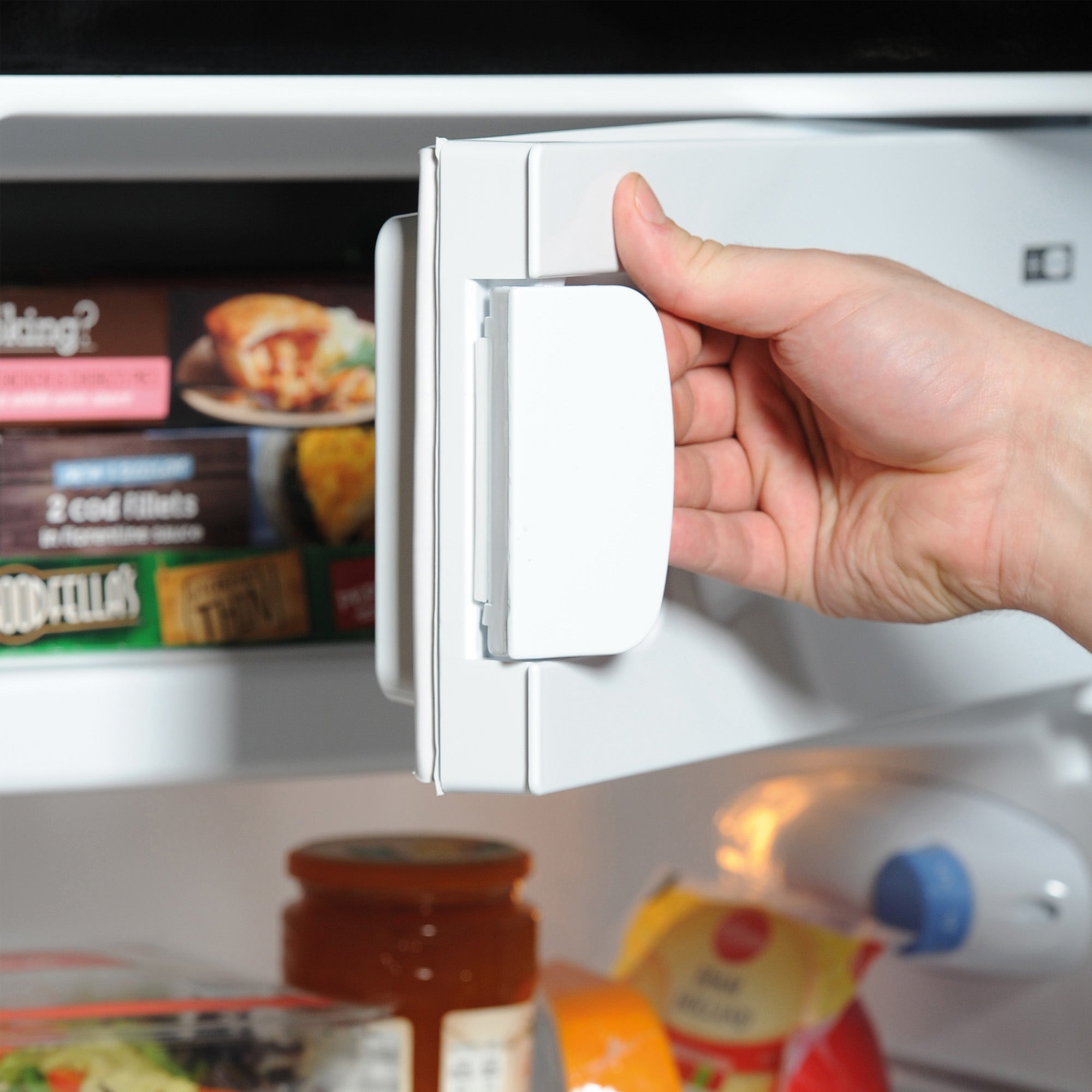 A hand opens the door of an Igenix Under Counter Fridge with Ice Box in black, revealing frozen food boxes, packaged items, and a jar on the lower shelves.