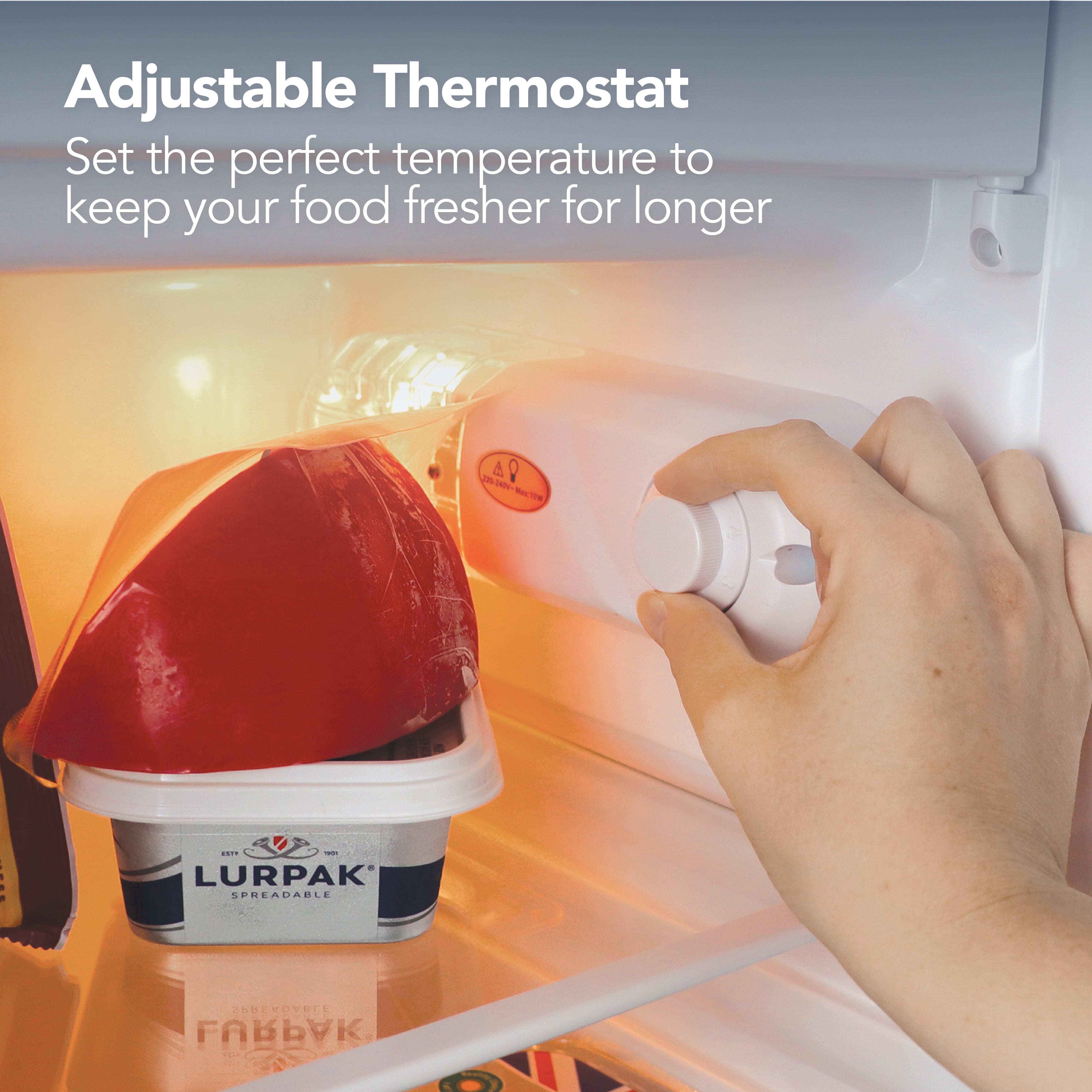 A hand adjusts the thermostat inside an Igenix Under Counter Fridge with Ice Box (81L, White) containing a red bell pepper and Lurpak Spreadable butter, highlighting how the perfect temperature keeps your food fresher for longer.