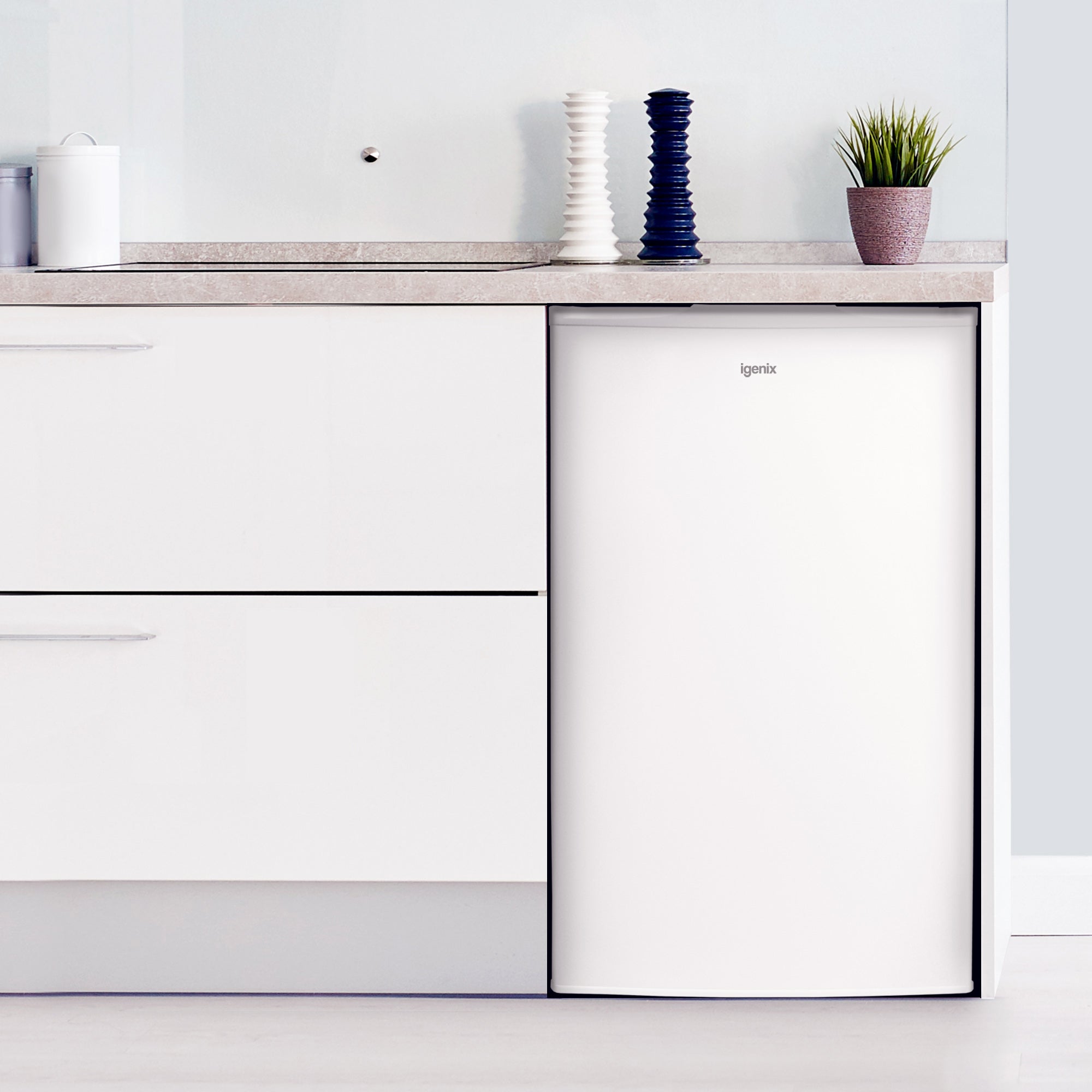 The Igenix Under Counter Fridge with Ice Box, 81 Litre, White, is seamlessly built into a modern kitchen beneath a light countertop. The bright, minimal space features a white canister, two tall textured vases, and a small potted plant.