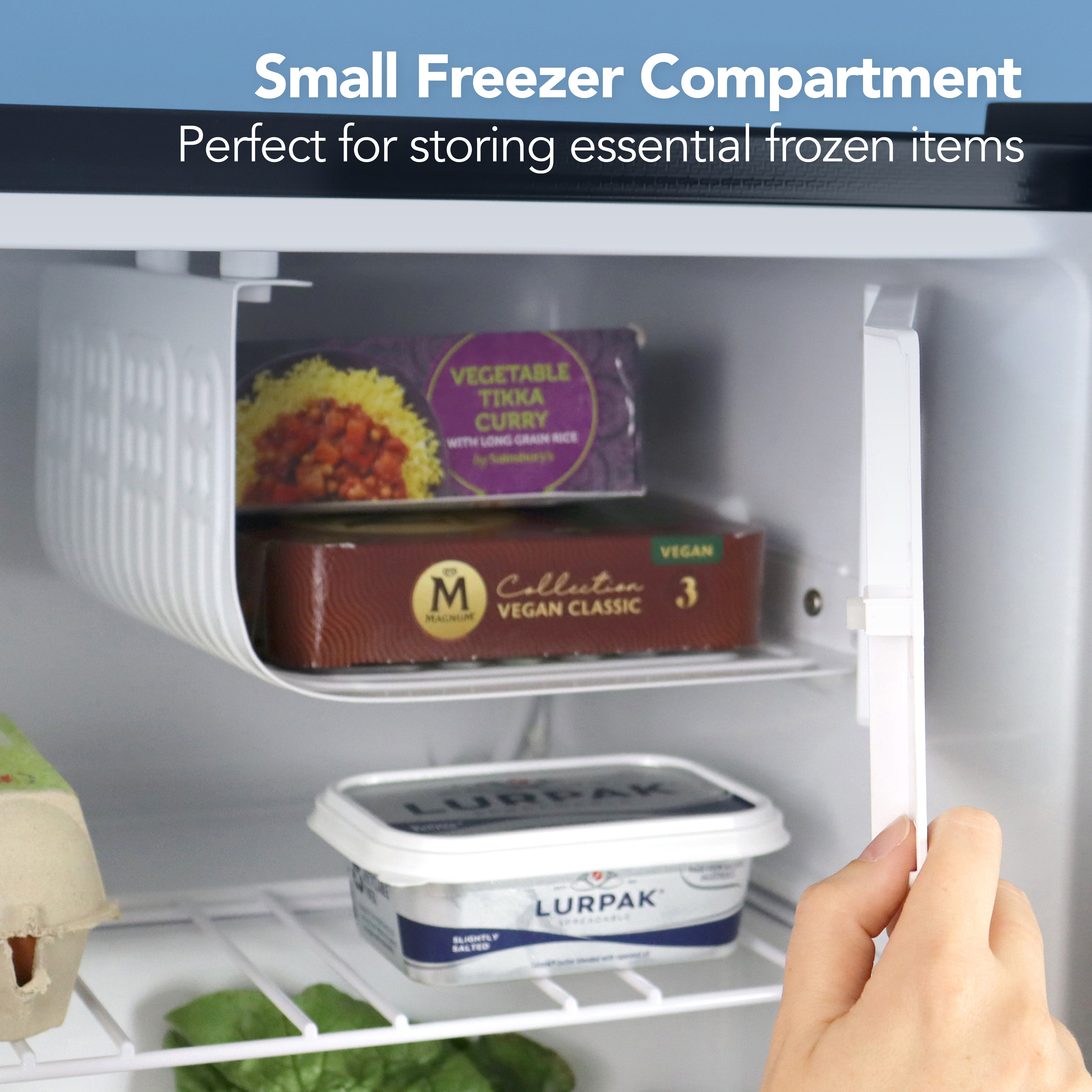 A hand opens the small freezer in the Igenix Table Top Fridge with Lockable Door, 41 Litre, Black—showing essentials like curry, vegan cheese, and butter. This compact fridge is ideal for small kitchens and perfect for storing frozen items.