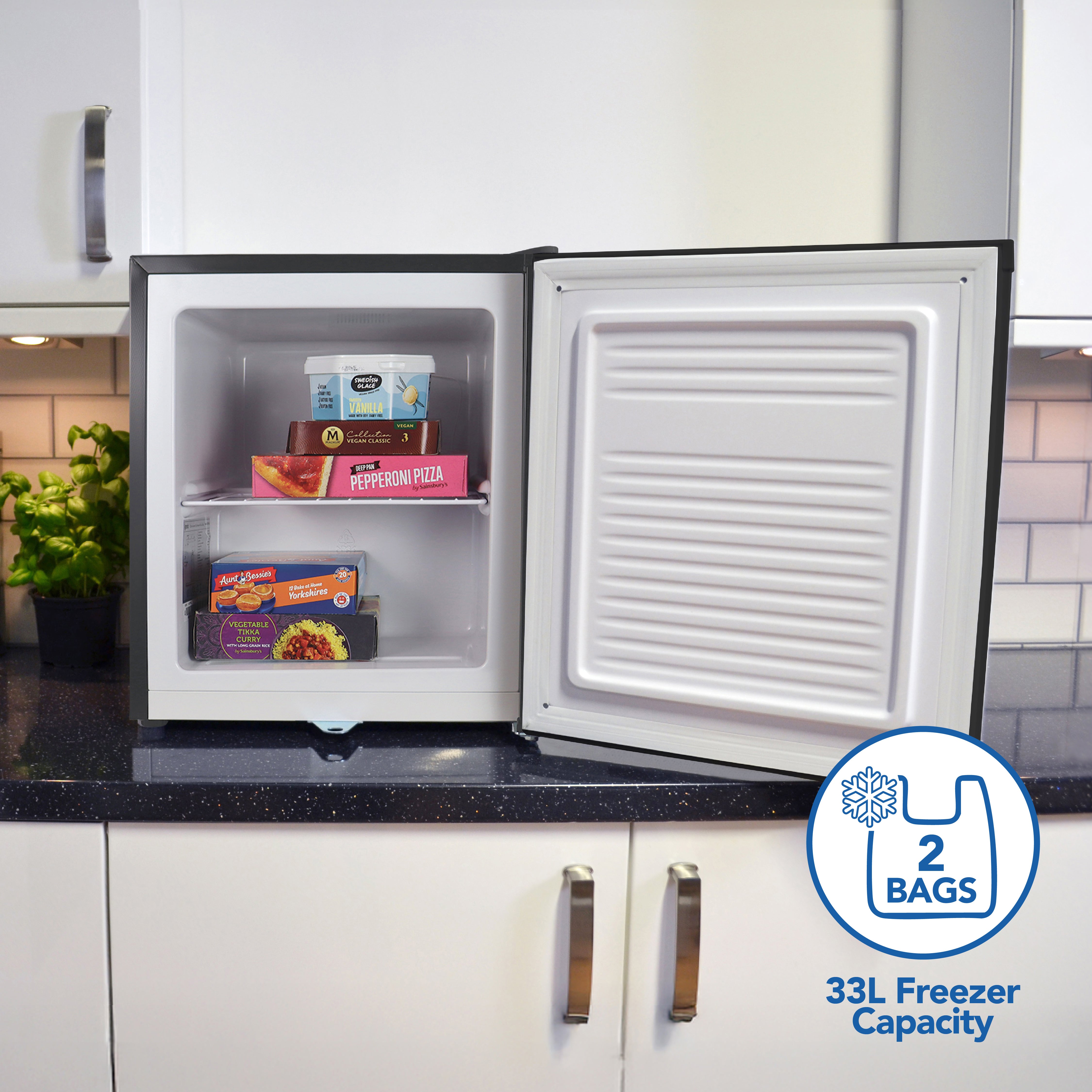 An Igenix Table Top Freezer with Lockable Door (33L, Black) sits open on a kitchen counter, holding frozen meals and pizza. A graphic overlay reads "2 bags, 33L Freezer Capacity." White cabinets and a basil plant are nearby.