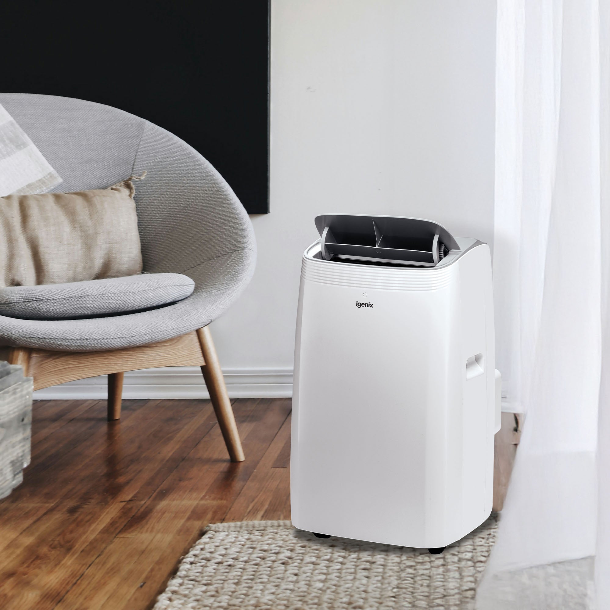 An Igenix 4-in-1 Portable Air Conditioner, 12000 BTU, is placed on a wooden floor beside a gray cushioned chair in a bright living room with sheer white curtains.