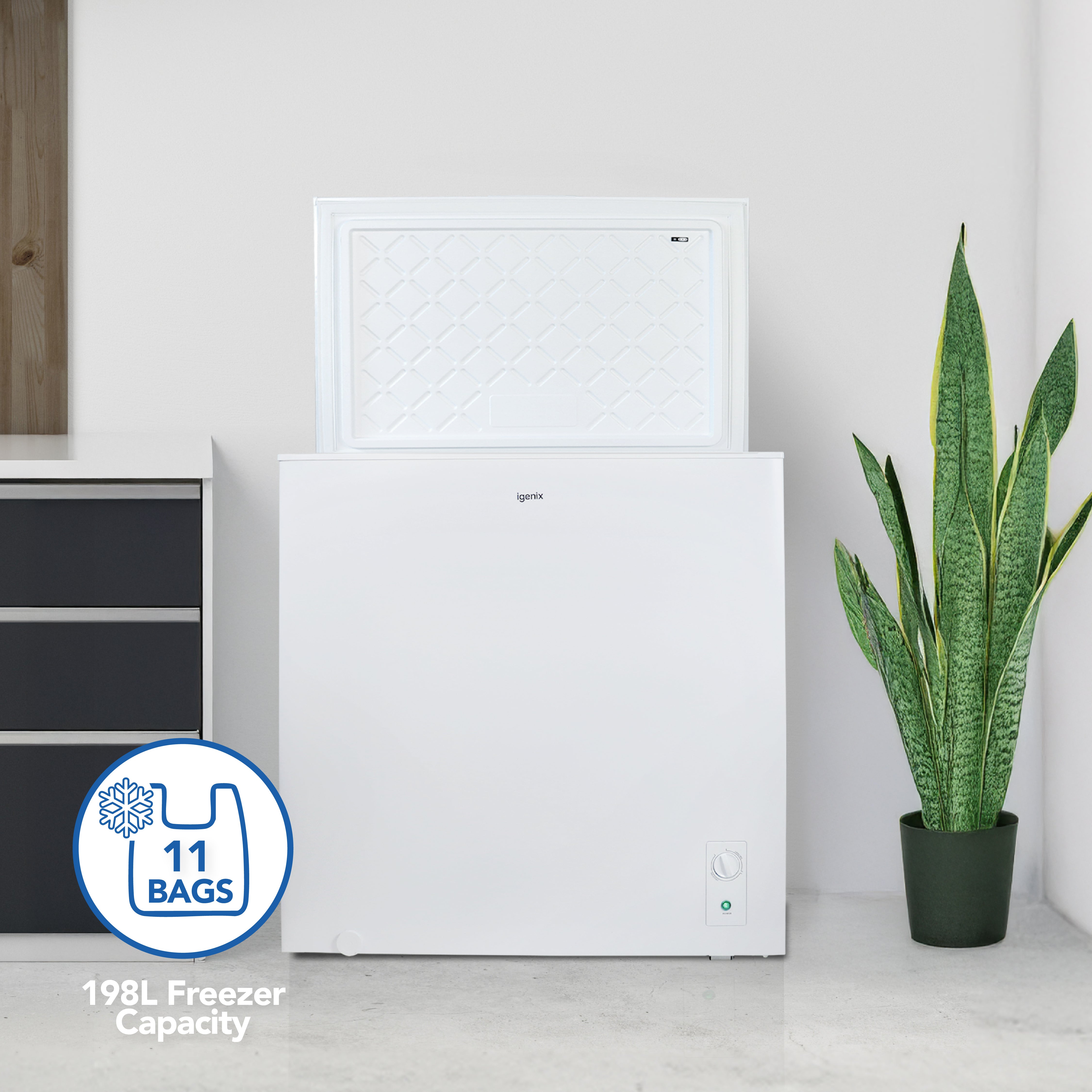 An Igenix Freestanding Chest Freezer, 198 Litre, White, stands beside a black cabinet and a tall green potted plant with its lid open. A graphic overlay highlights its 198L capacity and room for 11 bags.