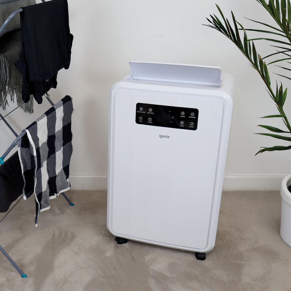 Igenix Portable Dehumidifier, Extracts 50 Litres per Day, White, with a digital control panel, standing on a carpeted floor next to a drying rack and a potted plant.