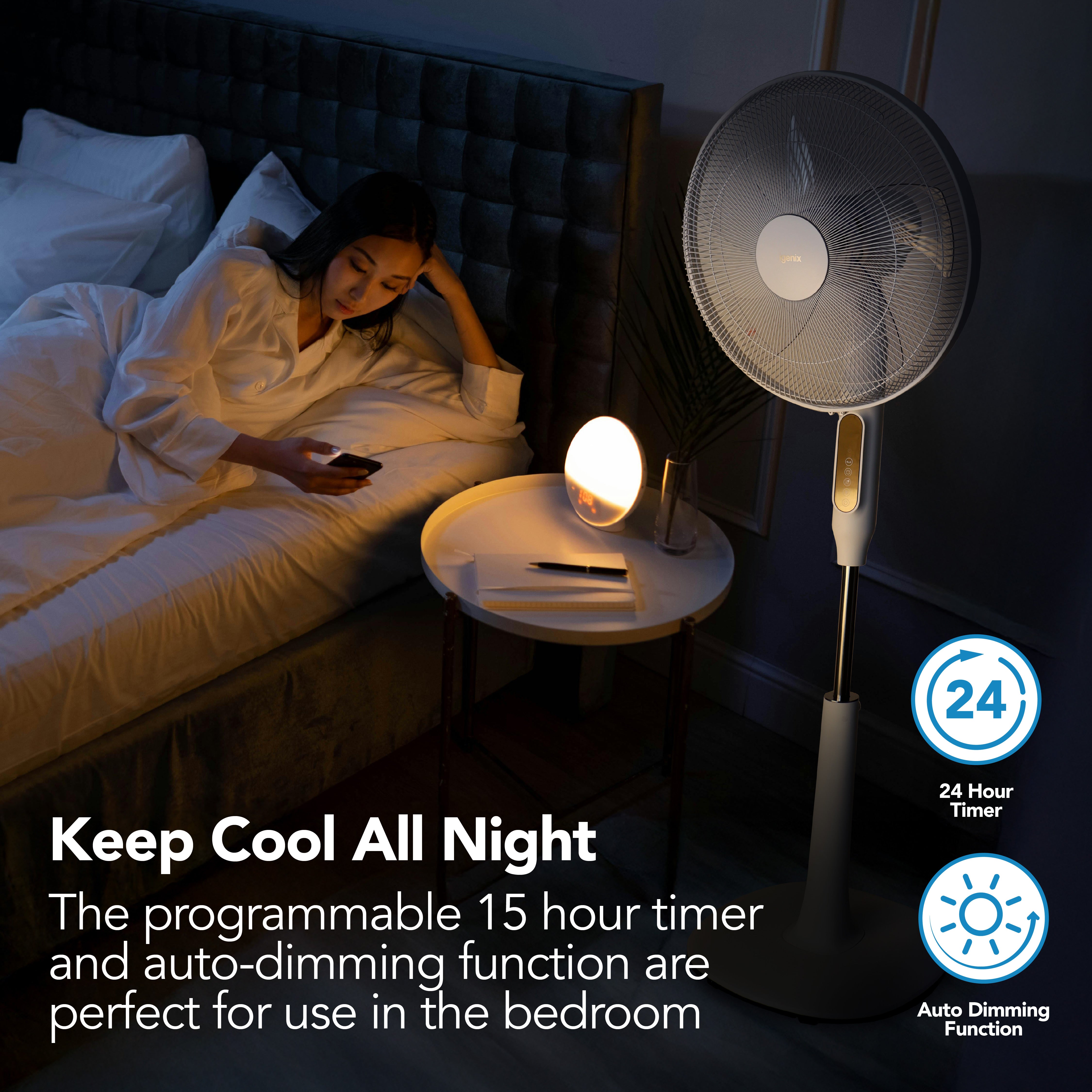 A woman sits on a bed using her phone beside a lit bedside lamp and an Igenix Digital Pedestal Fan. Text reads: "Stay cool all night—the programmable 15-hour timer and auto-dimming feature make it ideal for bedrooms.