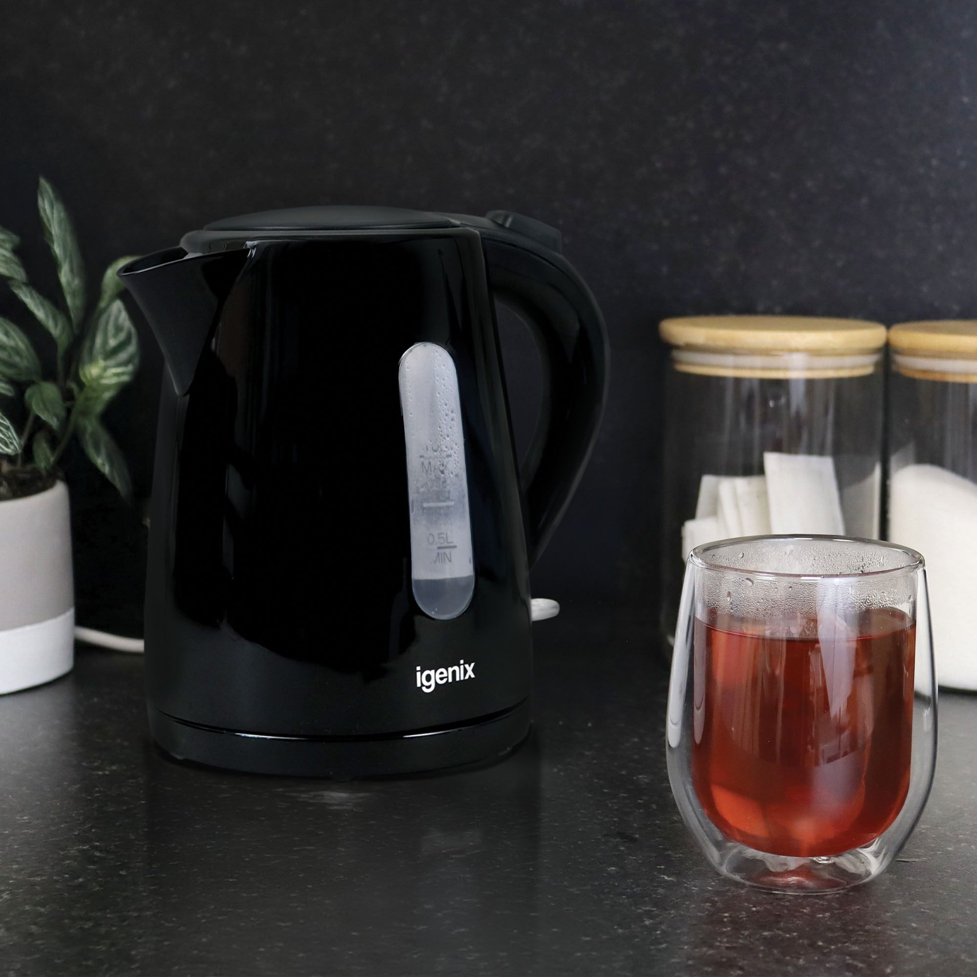An Igenix 1 Litre Cordless Jug Kettle, 2200W, Black, sits on a kitchen counter next to a glass of tea, a potted plant, and two jars containing white items—likely sugar and tea bags.