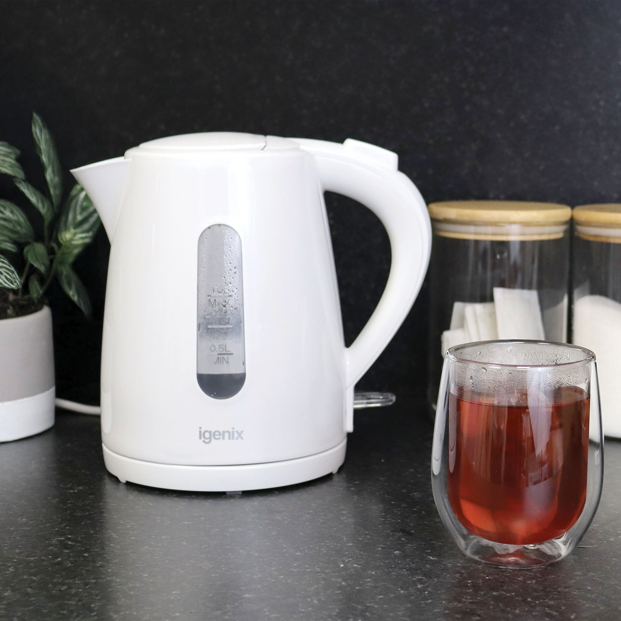 An Igenix 1 Litre Cordless Jug Kettle, 2200W in white sits on a dark countertop next to a glass of tea, two jars with wooden lids, and a small potted plant against a black background.