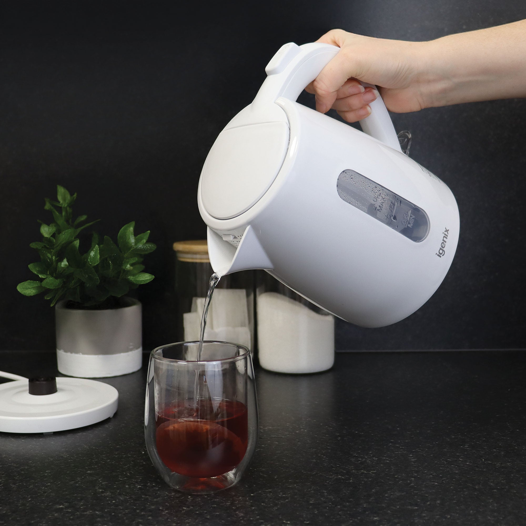 An individual pours hot water from the Igenix 1 Litre Cordless Jug Kettle, 2200W, White into a glass filled with red liquid. The scene unfolds on a dark kitchen counter adorned with a potted plant and jars in the background.