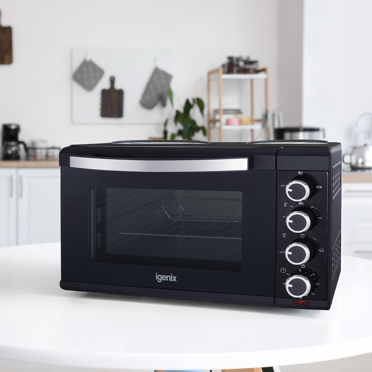 An Igenix 30L Mini Oven With Hotplates in black, featuring three control dials, sits on a white table in a modern kitchen with white cabinets and shelves in the background, showcasing a sleek and compact kitchen appliance designed for versatile cooking.
