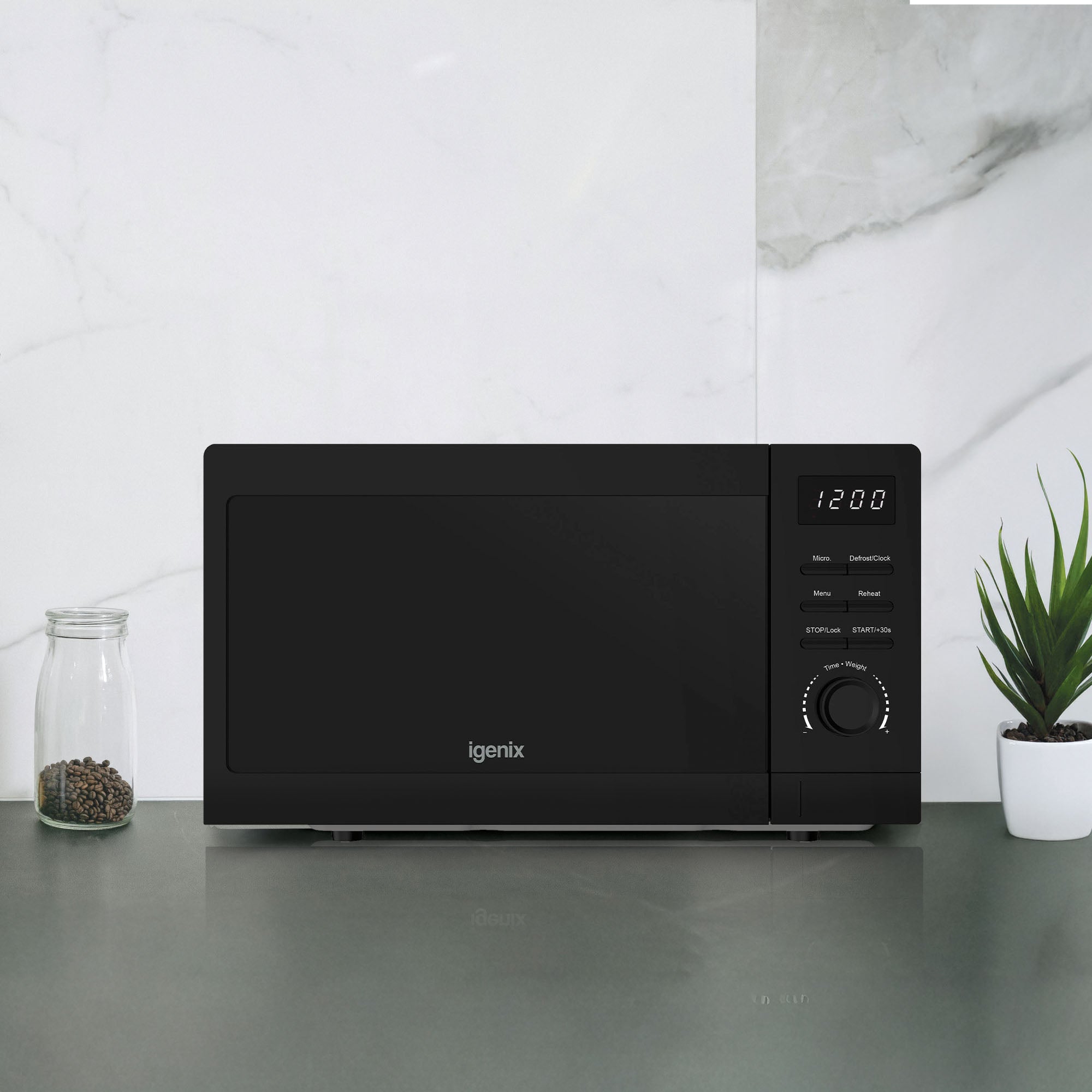 An Igenix IGMS02080B black 20L digital microwave with 5 power settings and 800W sits on a grey countertop, between a jar of peppercorns and a potted succulent, set against a white marble backsplash.