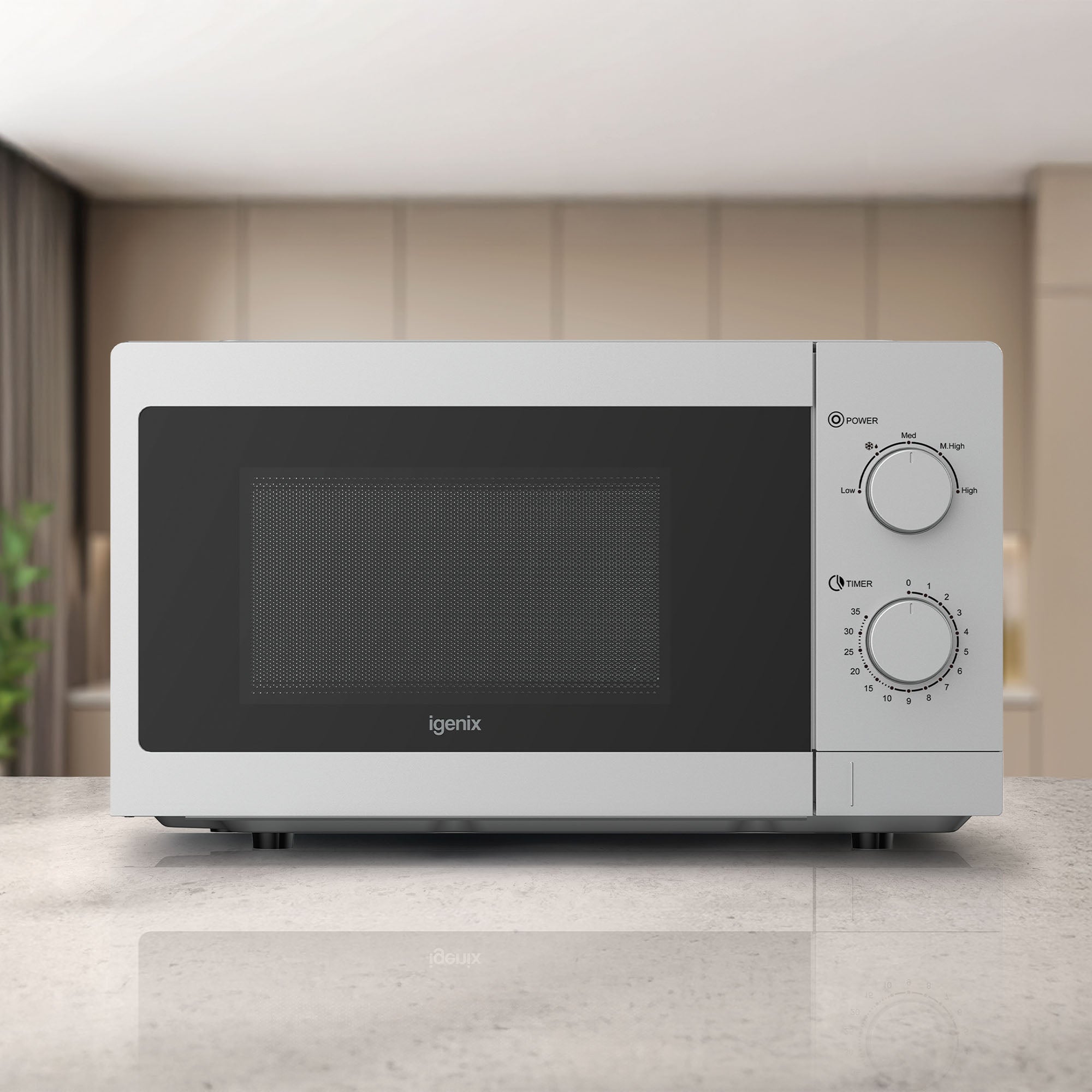 The Igenix Manual Microwave IGMS08080S, 20L, 800W in silver with 5 power settings and control dials sits on a kitchen counter, against blurred beige cabinets and a plant.