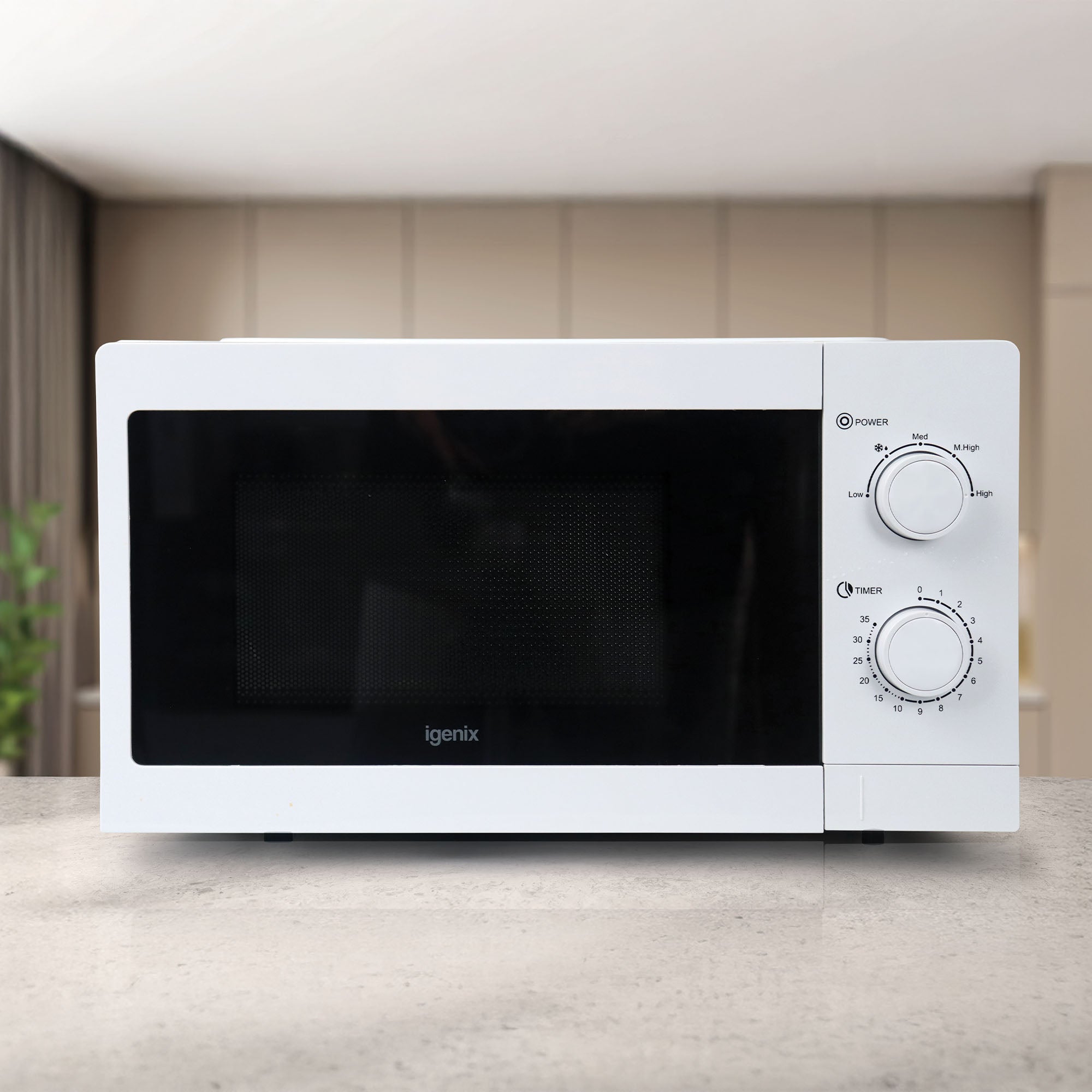 An Igenix Manual Microwave, 20L, white (IGMS08080W) with 5 power settings rests on a grey countertop in a modern kitchen with beige cabinets and a blurred green plant in the background.