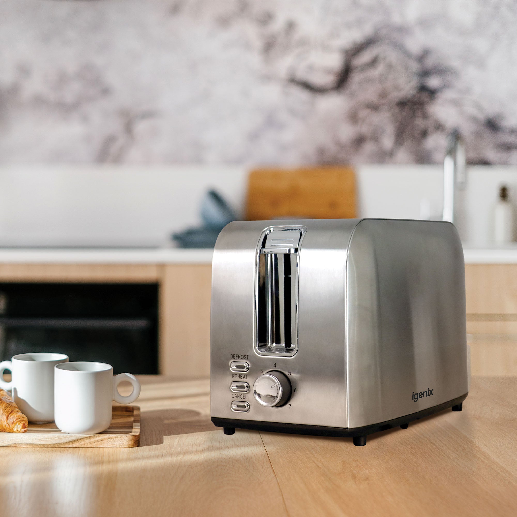 An Igenix 750W 2 Slice Toaster in stainless steel sits on a wooden kitchen counter with two white mugs and a croissant, set against a modern kitchen with light cabinets and a blurred backsplash.