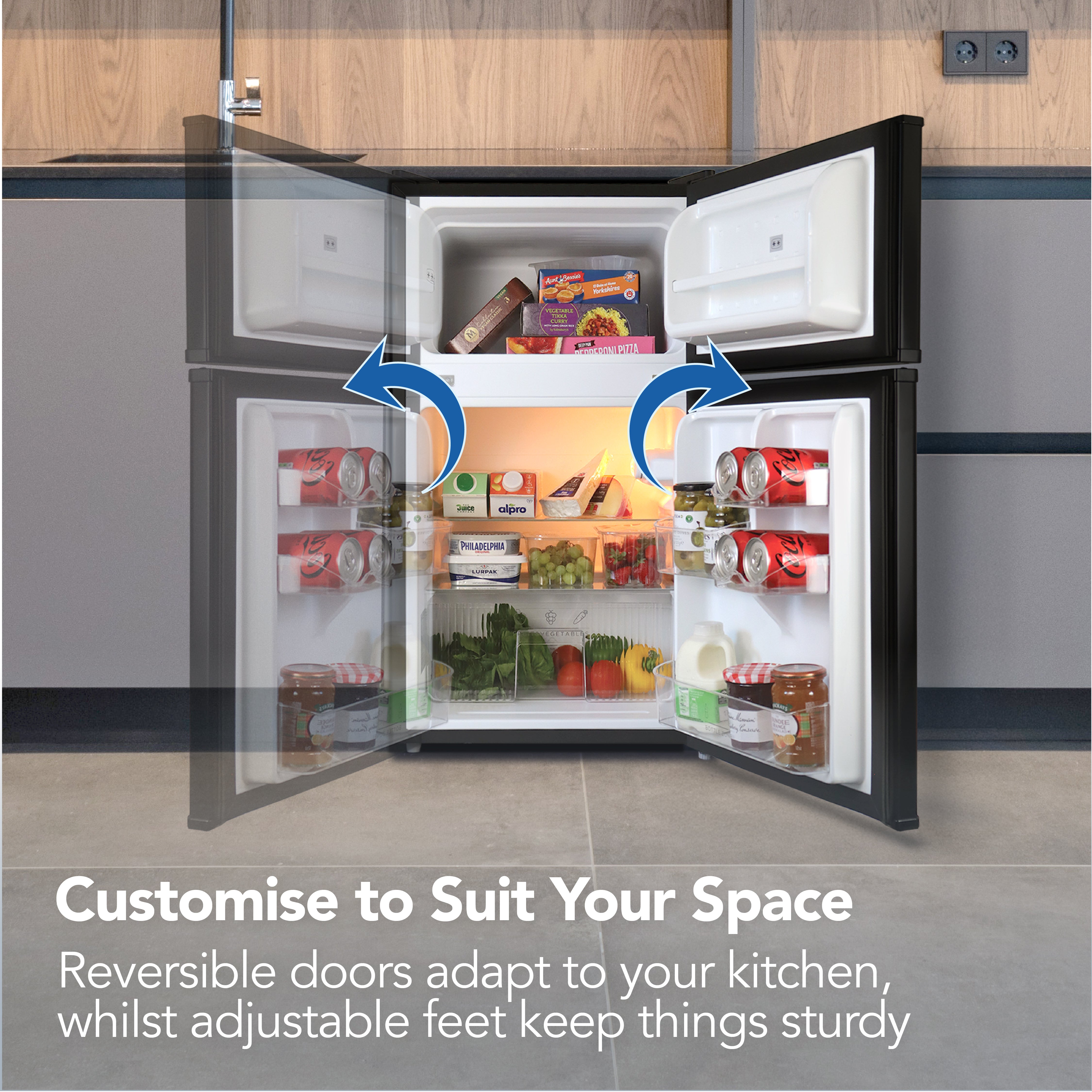 An open Igenix Under Counter Fridge Freezer (87L, Black) with food and drinks inside. Blue arrows on the doors highlight their reversible design. Text reads: Customise to Suit Your Space—features adjustable doors and feet.