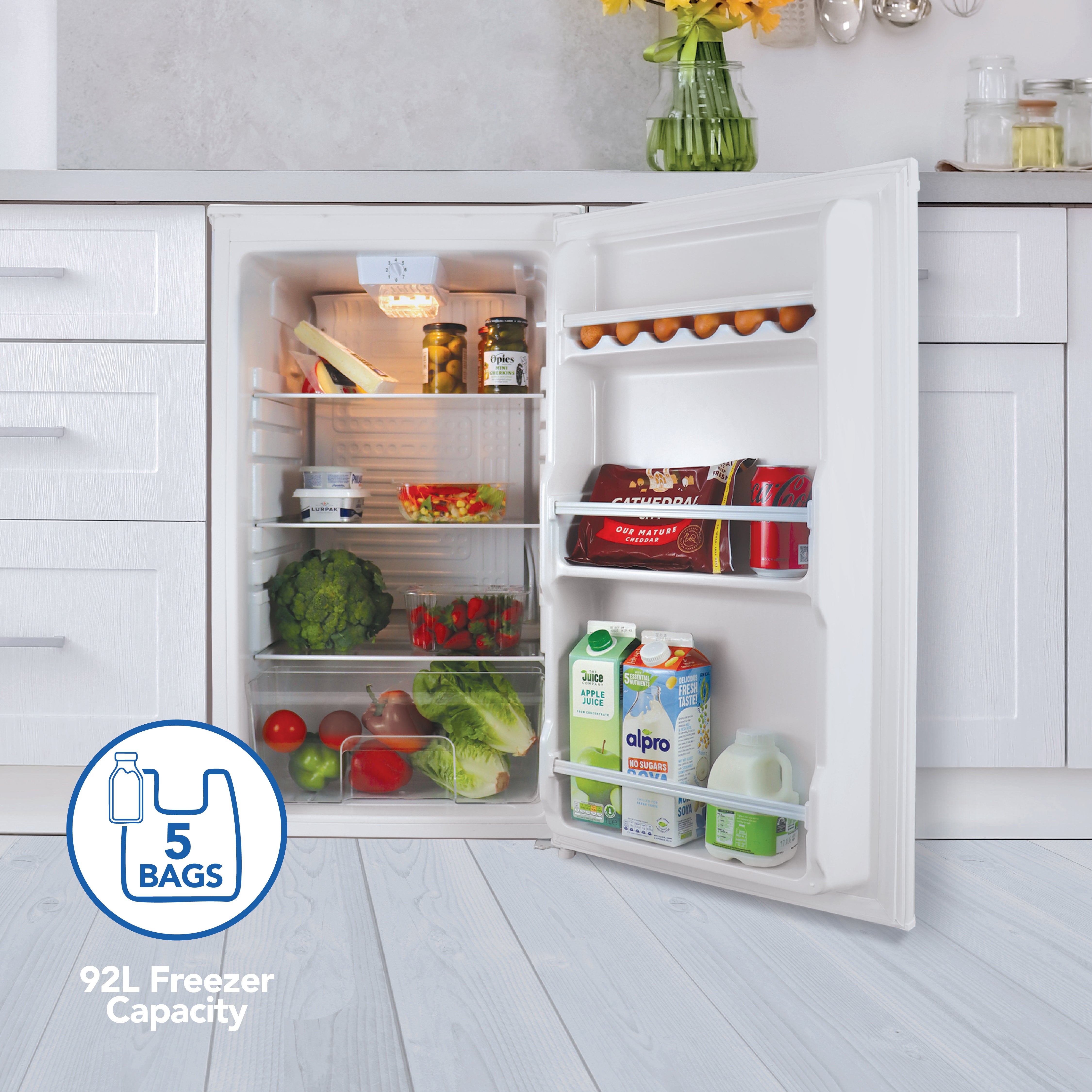 An open Igenix Under Counter Larder Fridge (92L, White) in a kitchen stores veggies, eggs, drinks, and snacks. A graphic shows its 92L capacity fits up to 5 bags—perfect for any compact kitchen fridge setup.