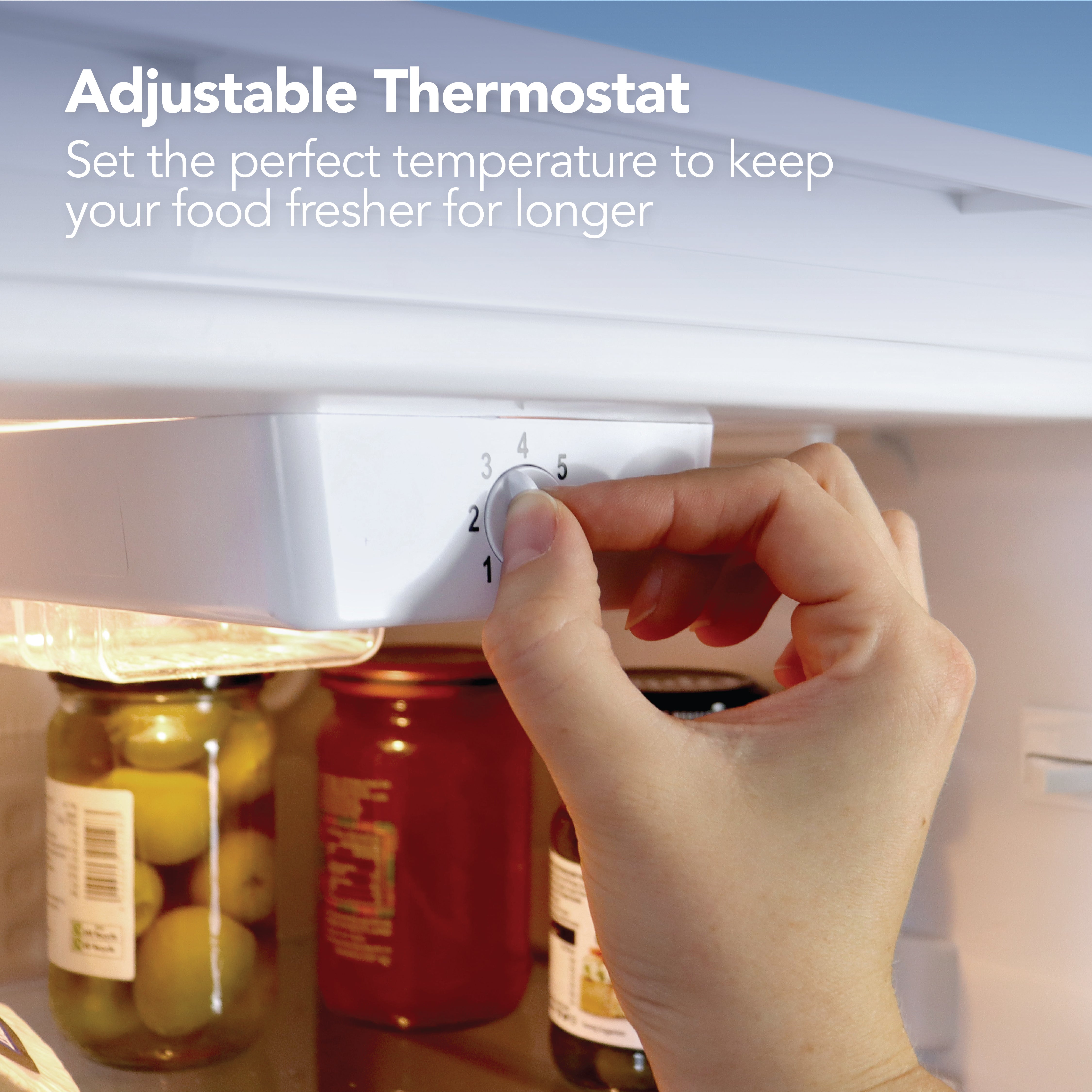 A hand adjusts the thermostat in the Igenix Under Counter Larder Fridge, 92L, White, with jars visible below. Text: Adjustable Thermostat—set the perfect temperature to keep your food fresher for longer.