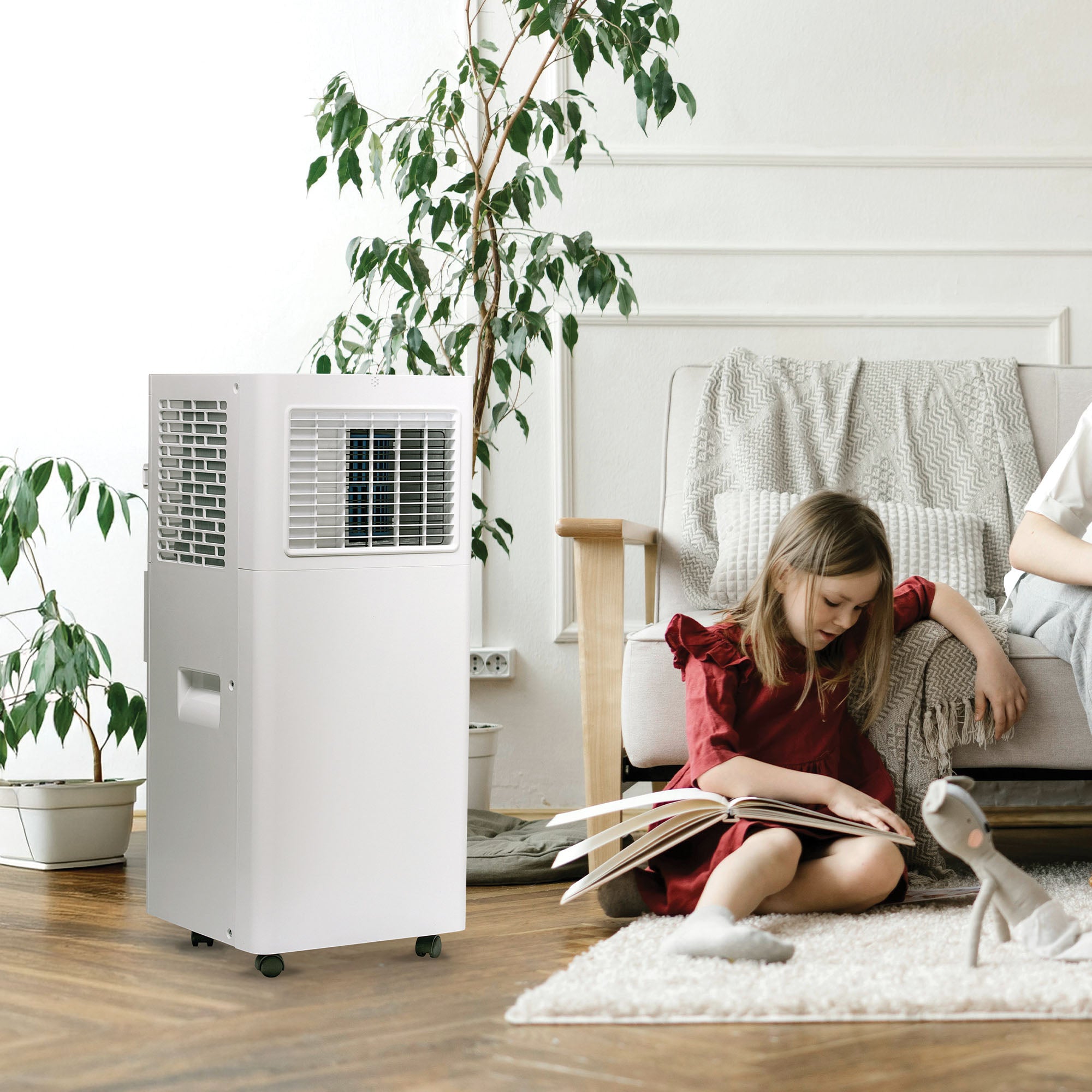 A young girl in a red dress sits on the floor reading near an Igenix 3-in-1 Portable Air Conditioner (Cooling, Fan & Dehumidifier, 9000 BTU) in a bright, cozy living room with a sofa, blanket, potted plant, and plush toy.