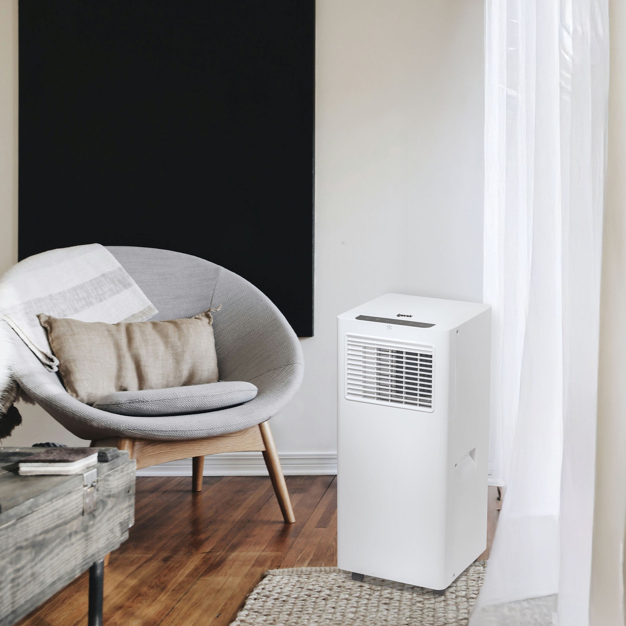A modern living room with a gray rounded armchair beside a small table, light wood flooring, and the Igenix 3-in-1 Smart Air Conditioner (9000 BTU) near sheer white curtains and bold large black wall art.