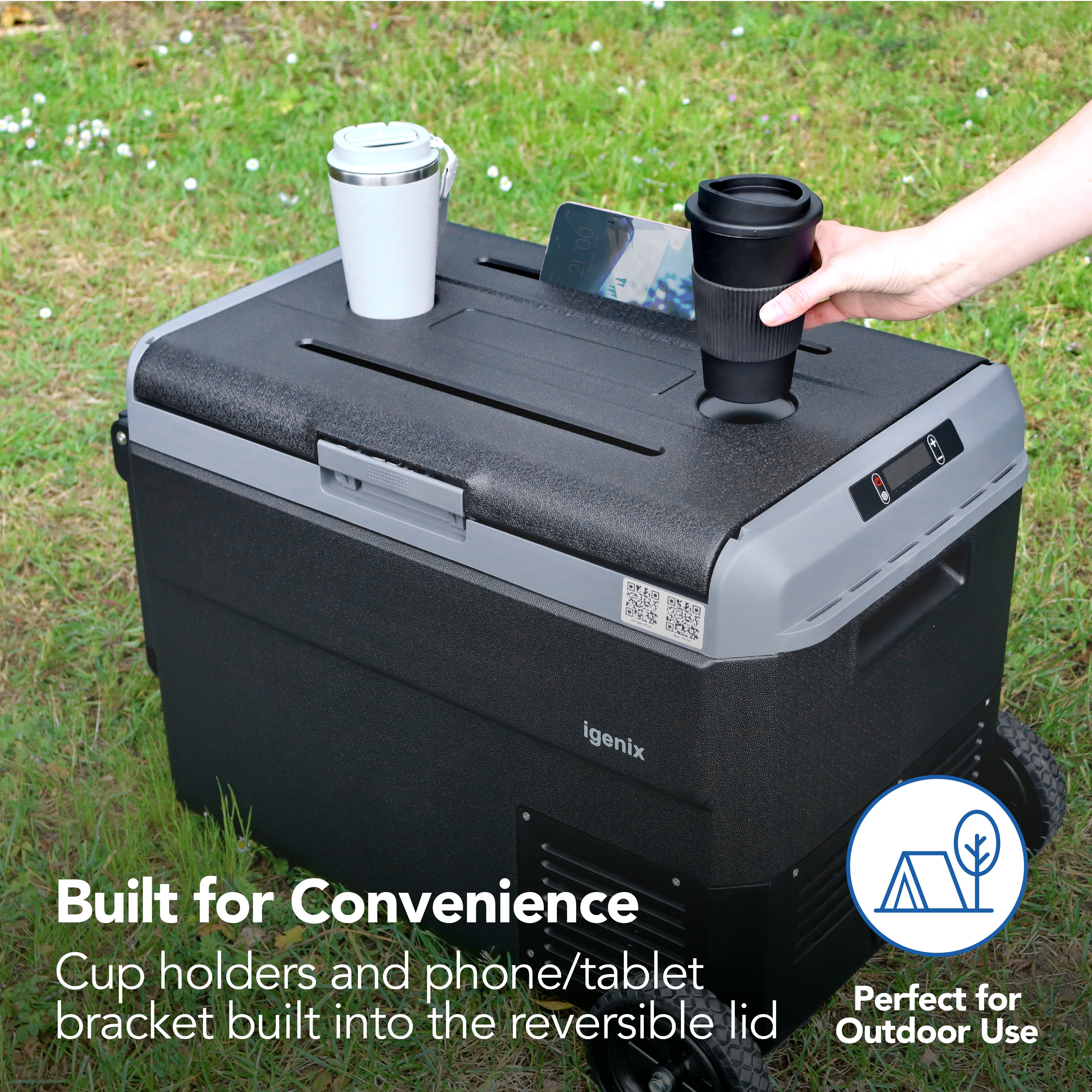The Igenix 50L Portable Fridge Freezer in black sits on grass, featuring cup holders and a phone/tablet bracket on its lid—shown holding two travel cups and a phone. Text: Built for Convenience and Perfect for Outdoor Use.