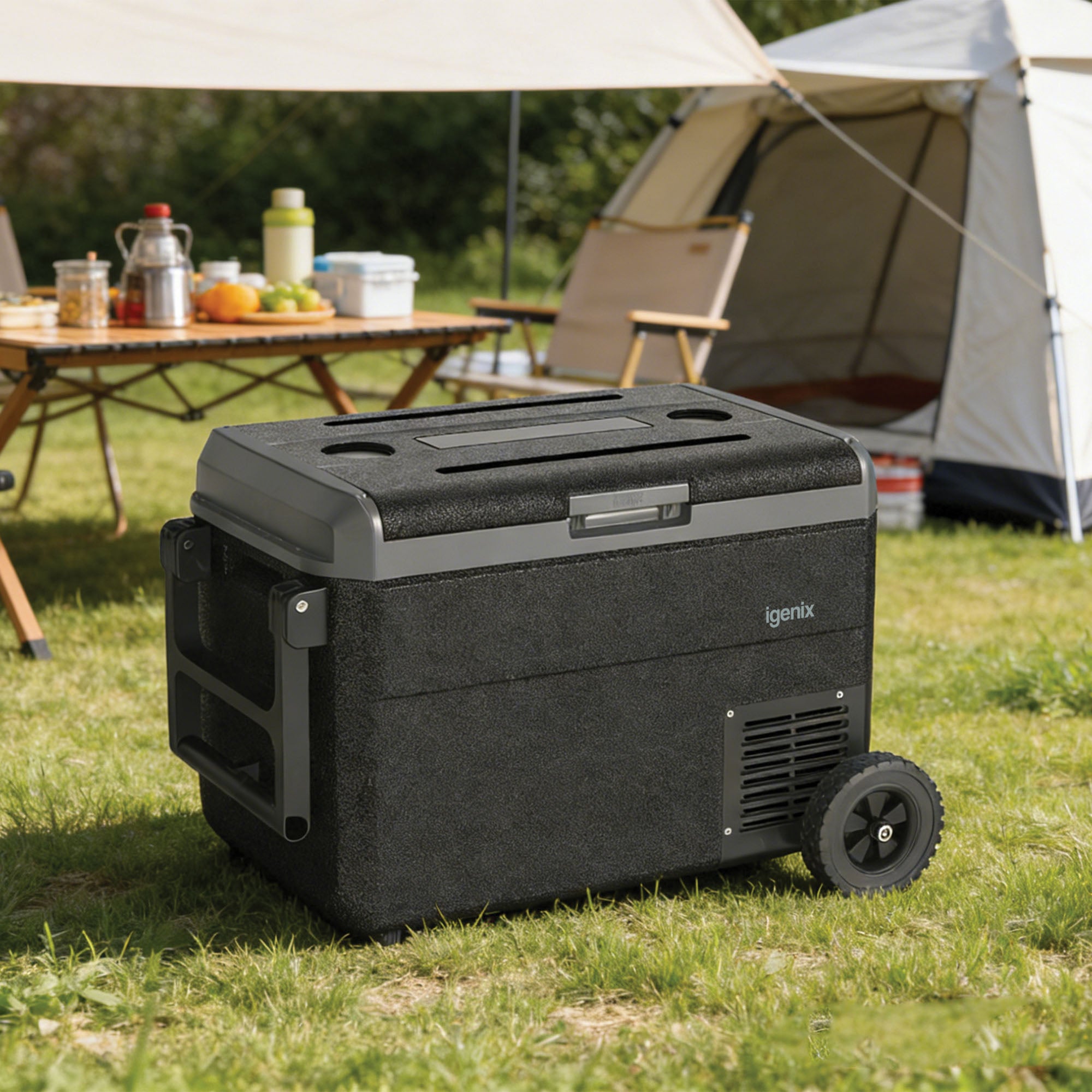 An Igenix 50L Portable Fridge Freezer in black is placed on grass at a campsite, with a tent and a wooden table featuring food and drinks visible in the background.