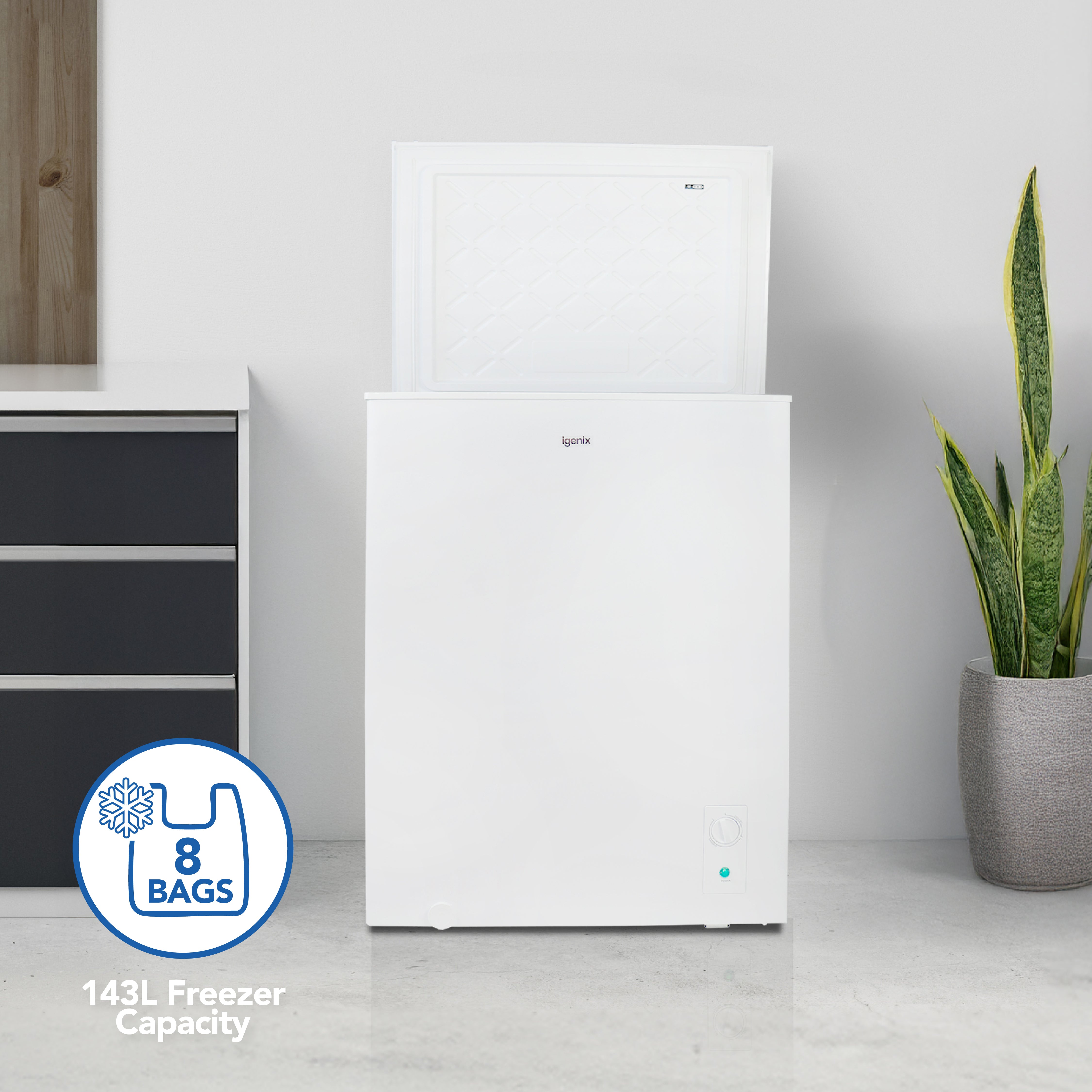 The Igenix Freestanding Chest Freezer (143L, White) stands open on a gray floor by a plant and black cabinet. Its spacious 143-litre capacity holds 8 bags, making this energy-efficient freezer ideal for extra storage.