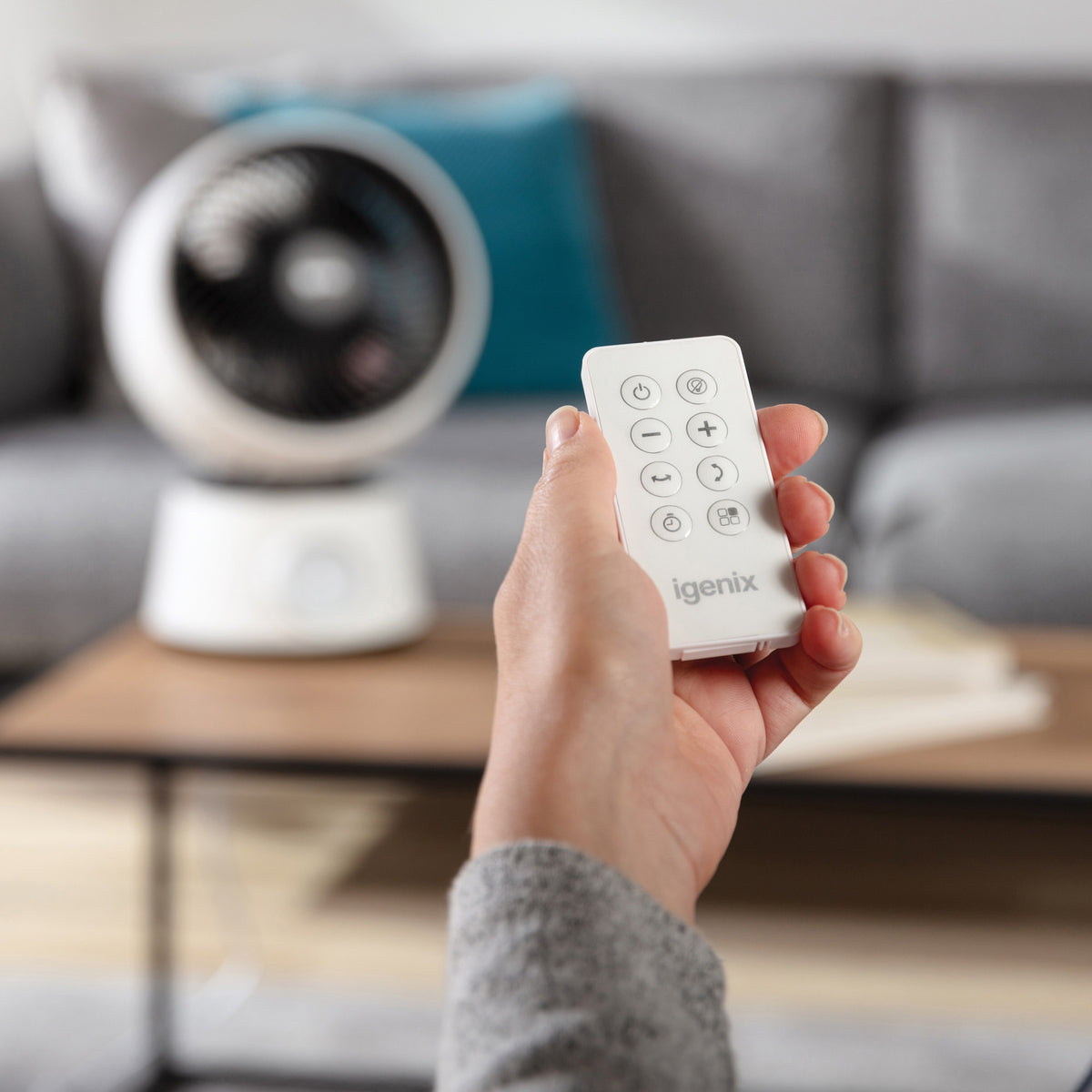 A hand holds a white remote control with circular buttons, pointed toward an Igenix Digital Air Circulator Turbo Fan, 9 Inch, White sitting on a coffee table.