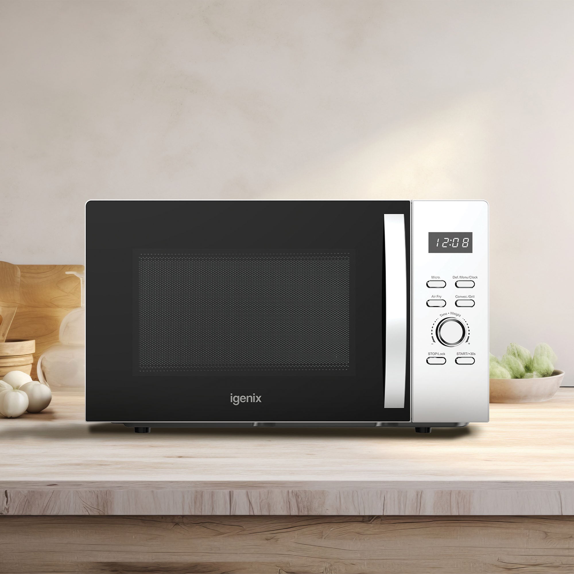 A white Igenix Digital Microwave (25L, 60 min timer, air fryer, 850W, IGMC02580W) is on a wooden kitchen counter; its digital display shows 12:08 with vegetables and kitchen items in the background.