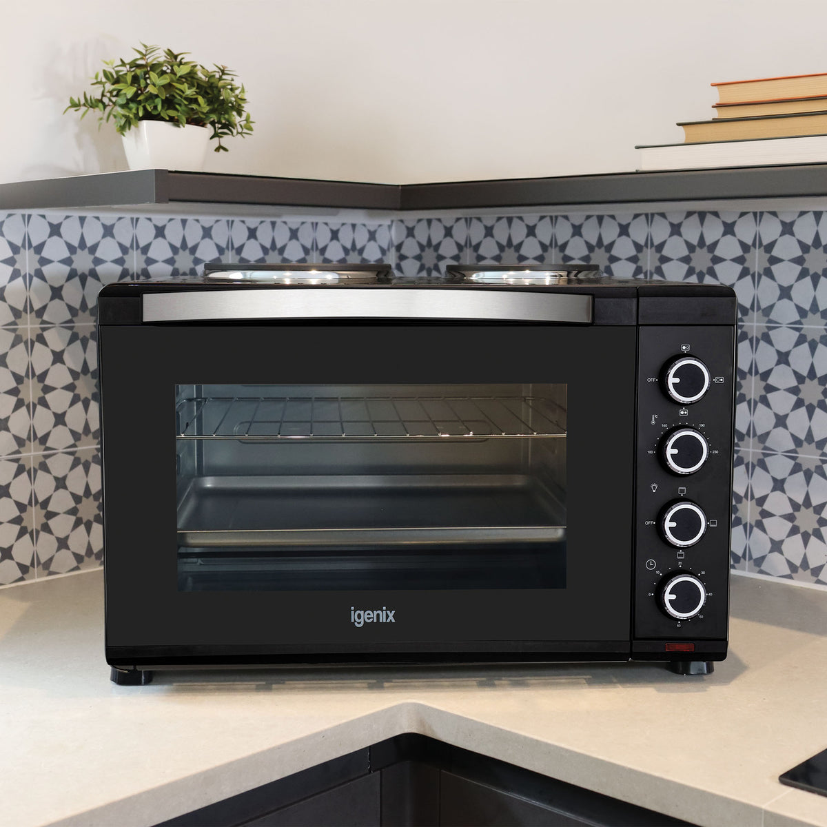 An Igenix 30L Mini Oven With Hotplates in black, featuring four control knobs and convenient double hotplates, sits on a kitchen counter adorned with patterned blue and white tiles and a small plant in the corner.