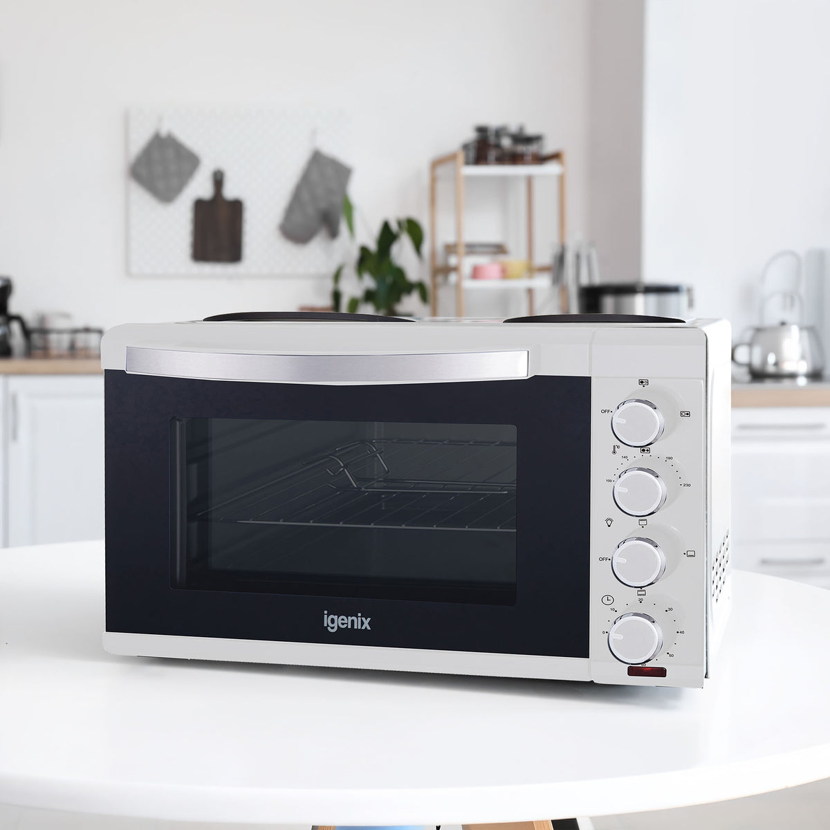 A white Igenix 30L Mini Oven With Hotplates is placed on a pristine kitchen counter. This compact kitchen appliance, equipped with control dials and double hotplates, blends seamlessly into the modern setting where shelves and household items are neatly arranged in the background.