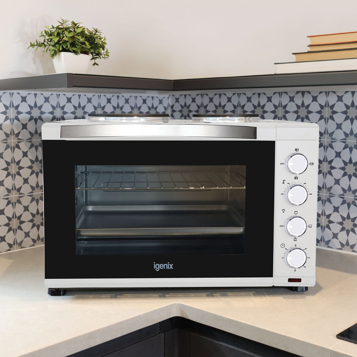 An Igenix 30L Mini Oven With Hotplates, White featuring control knobs and double hotplates sits in a kitchen corner, set against a patterned blue and white tile backsplash.