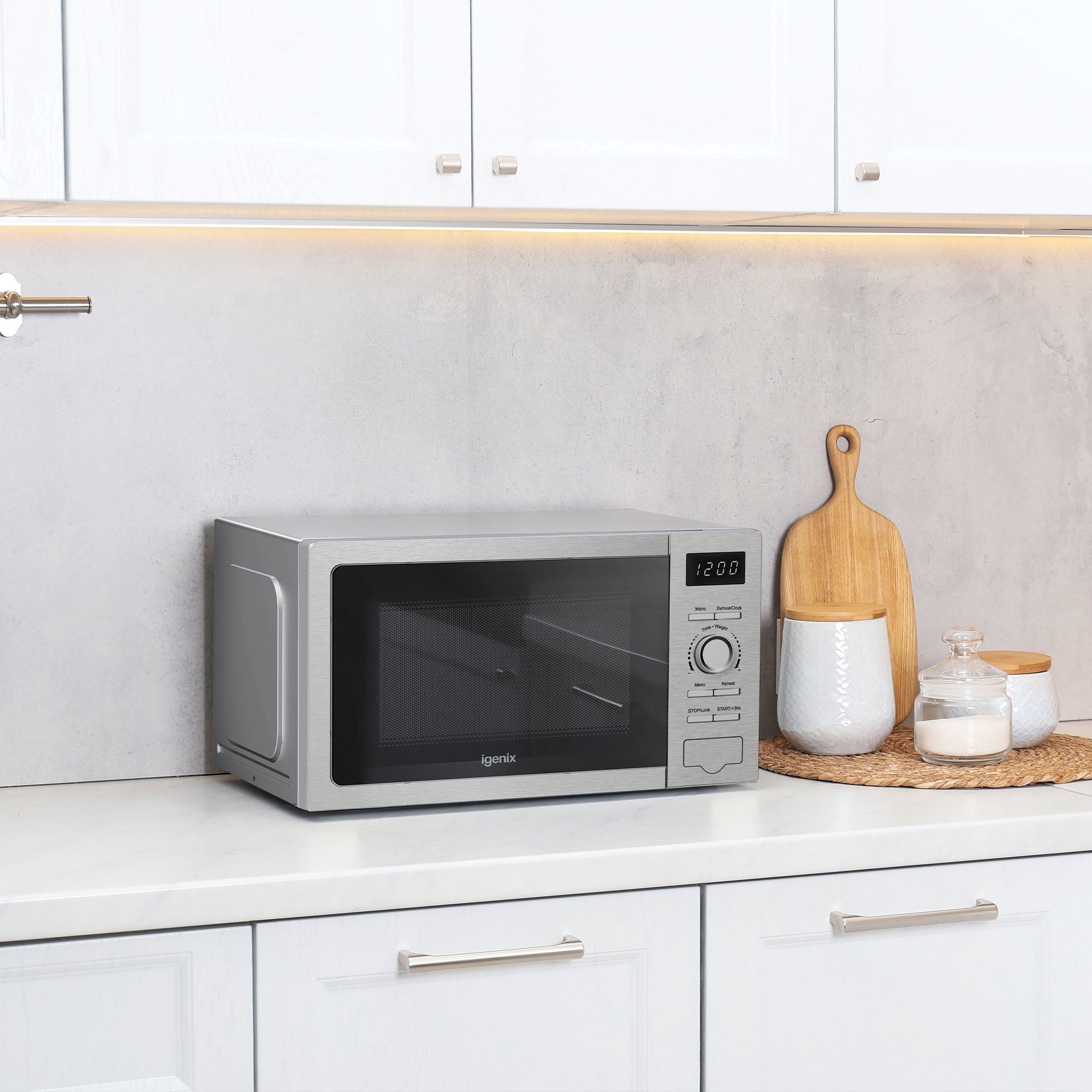An Igenix Digital Microwave (20 Litre, 5 Power Settings, 800W, Stainless Steel, IGMS02080SS) sits on a white kitchen counter beneath cabinets, next to a cutting board, a jar, a basket, and a ceramic container. The microwave display shows 12:00.