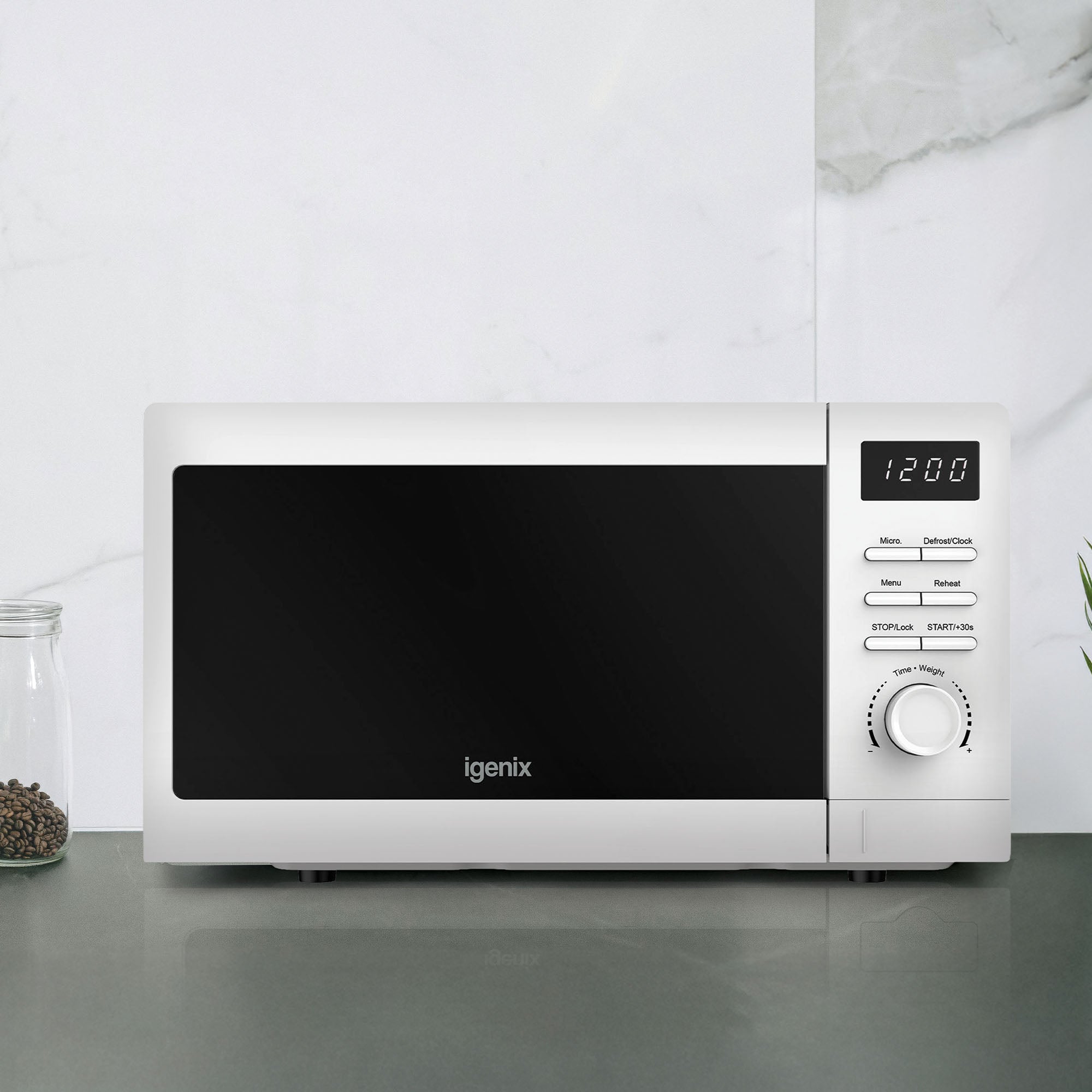 An Igenix Digital Microwave, 20L, 800W, white (IGMS02080W) with a clock display at 12:00, rotary dial and buttons sits on a grey countertop beside two glass jars against a white marble-patterned wall.