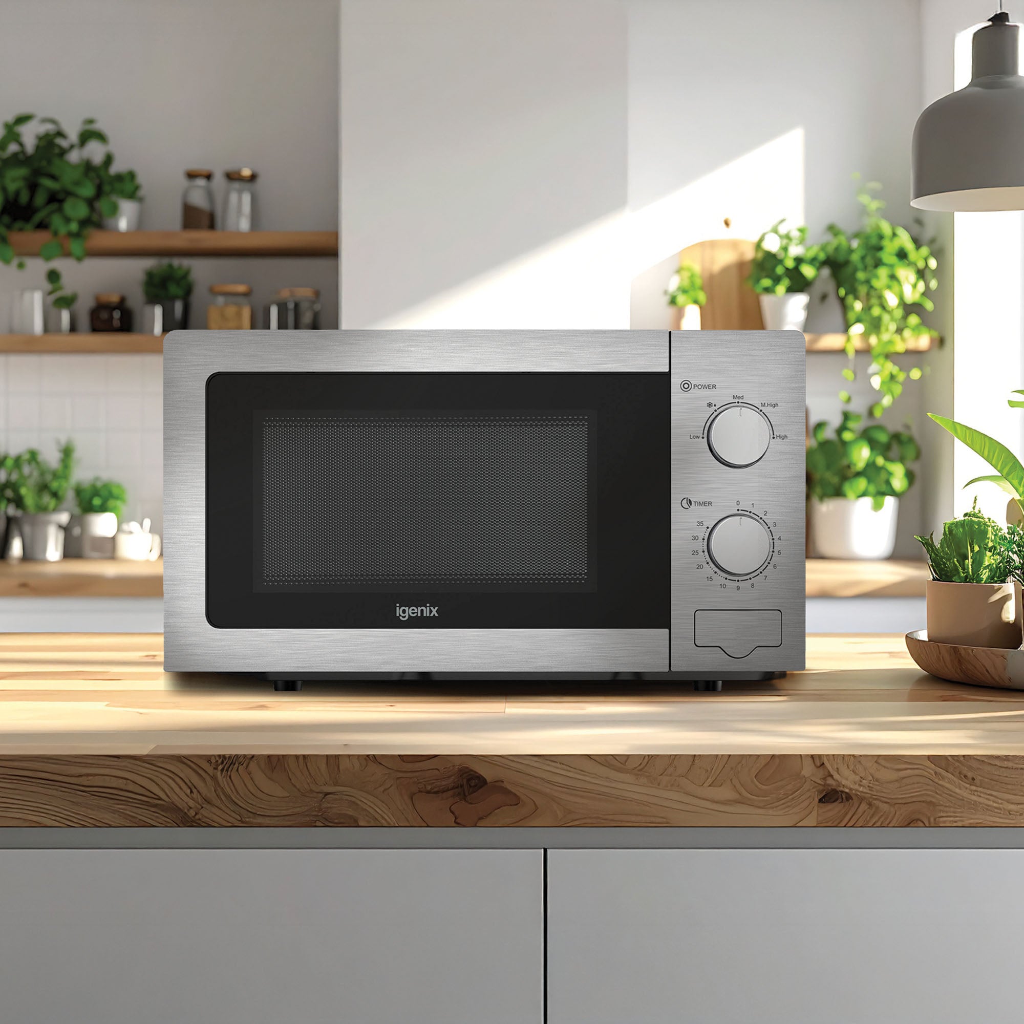 An Igenix Manual Microwave, 20L, 5 Power Settings, 800W (IGMS02081SS) in stainless steel sits on a wooden kitchen countertop with sunlight streaming in. Modern shelves behind hold plants and dishes in a tidy kitchen.