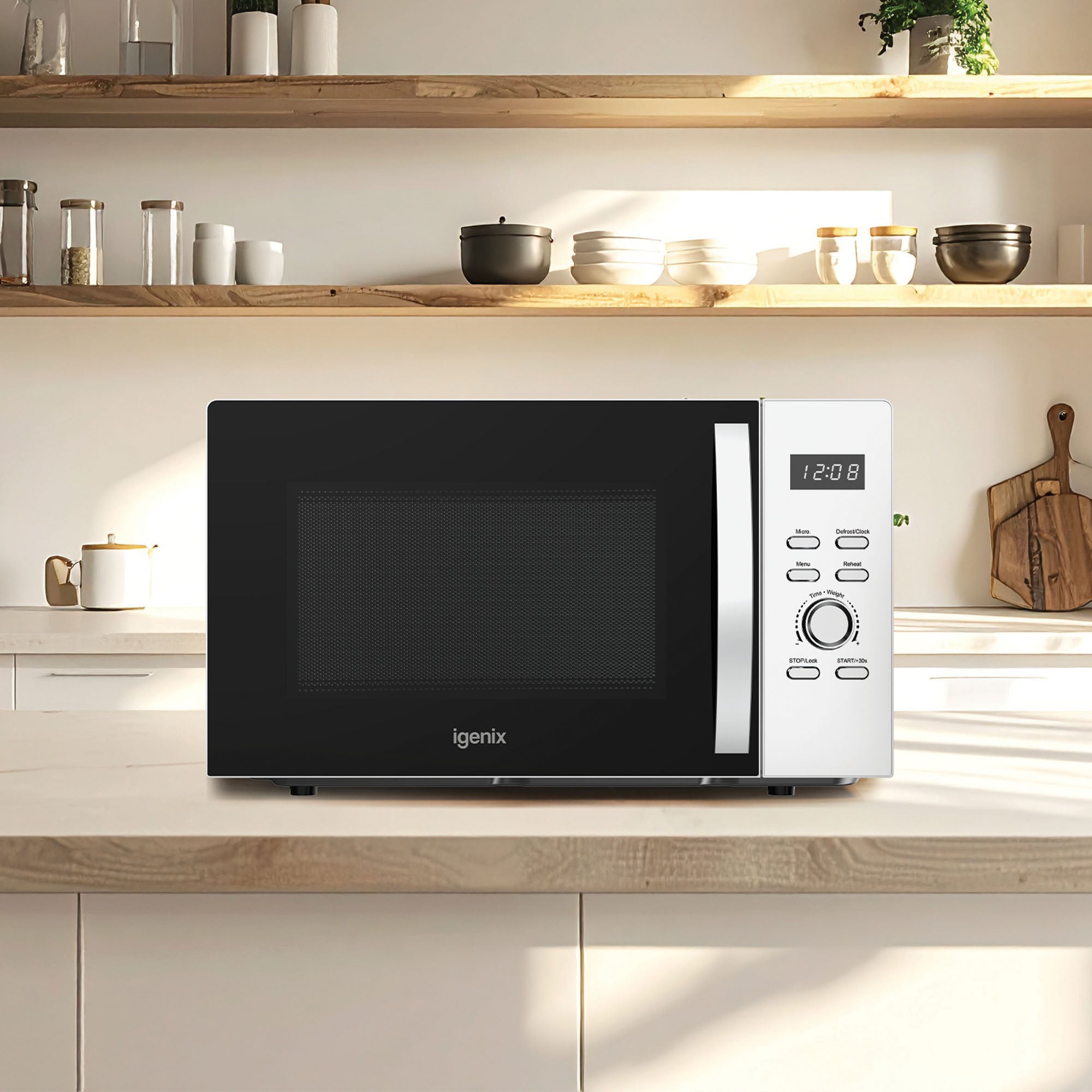 An Igenix IGMS03090W 30L 950W digital microwave in white sits on a kitchen counter, beneath wooden shelves holding bowls, jars, and cups in a bright, minimalist kitchen.