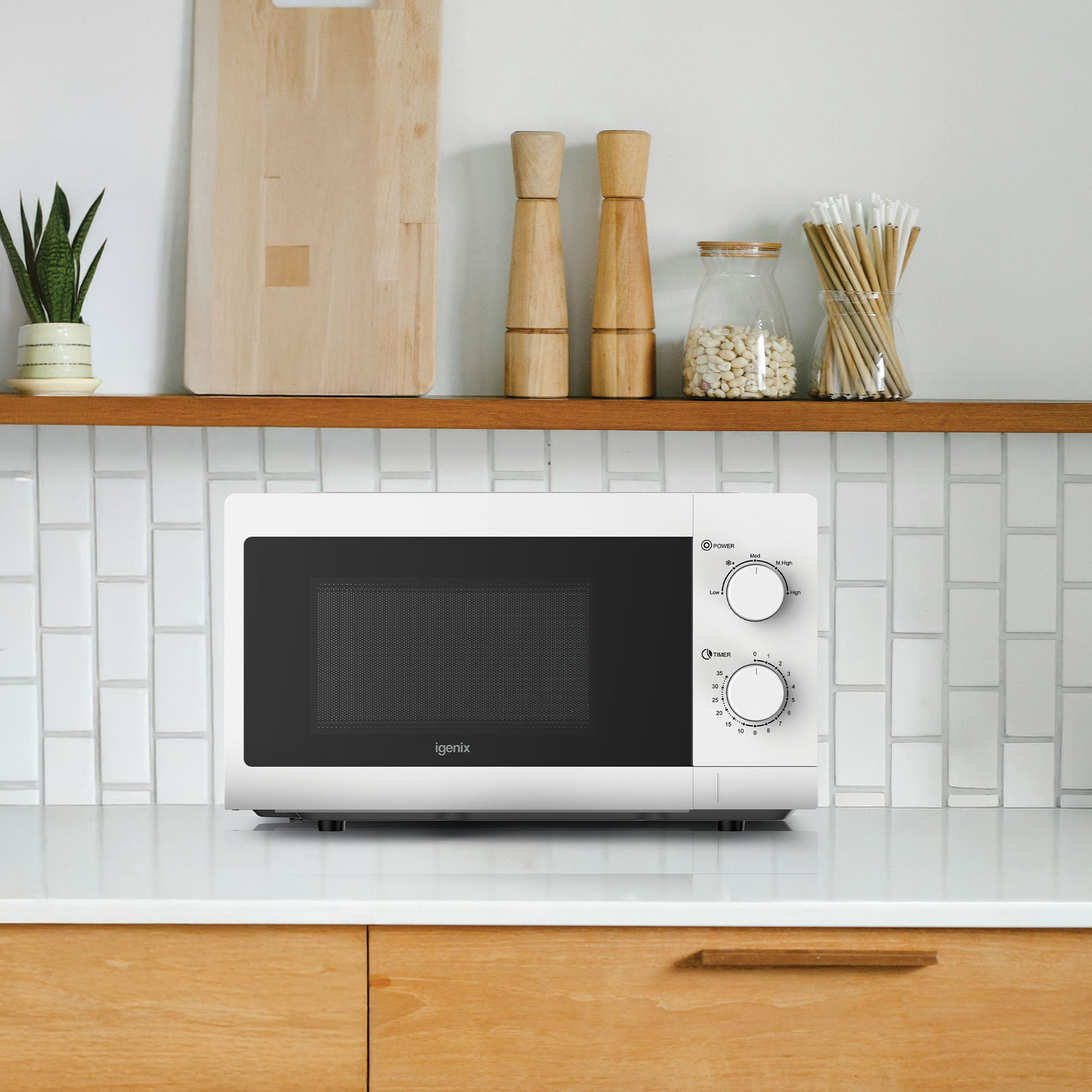 An Igenix Manual Microwave, 20L, 700W, IGMS09434W in white with two dials sits on a white countertop in a modern kitchen, alongside a tiled backsplash, plant, wooden utensils, and jars.