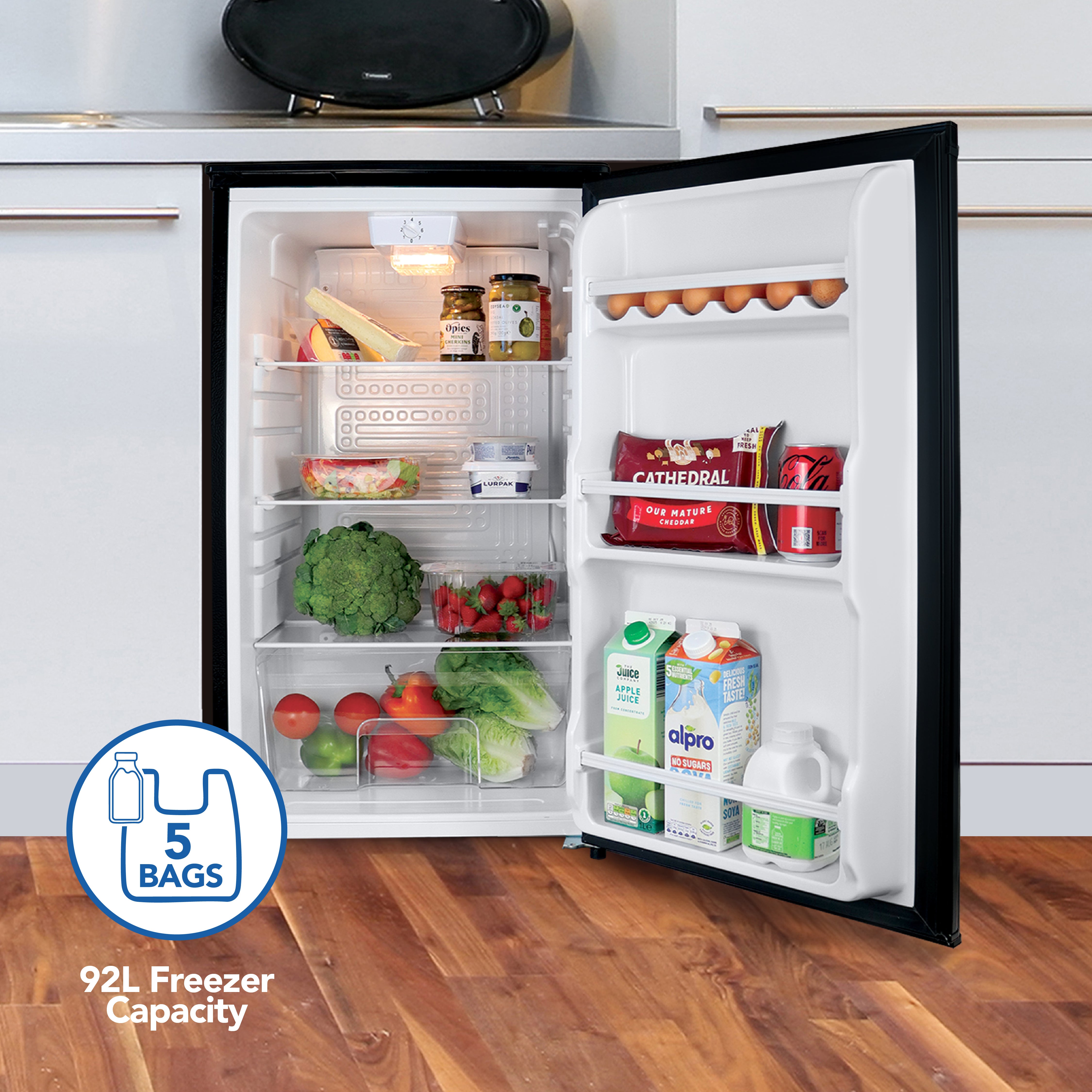 The Igenix Under Counter Larder Fridge (92L, reversible door, black) stands in a modern kitchen with wooden floors. Inside are eggs, milk, juice, cheese, vegetables, and strawberries. A badge notes capacity for 5 bags.