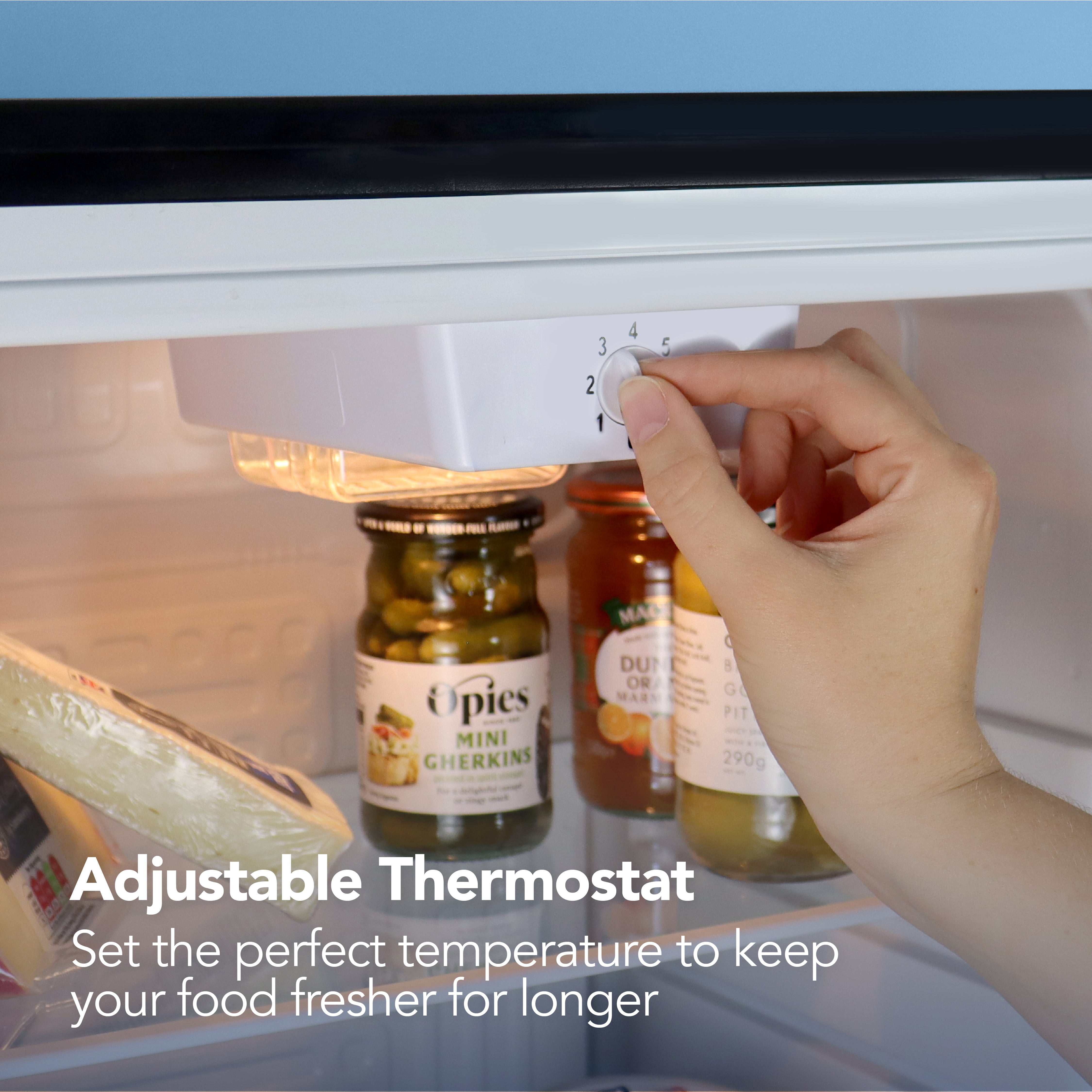 A hand adjusts the thermostat dial inside the Igenix 92 Litre Under Counter Larder Fridge, with jars and cheese on the shelf. Text reads: Adjustable Thermostat – Set the perfect temperature to keep your food fresher for longer.