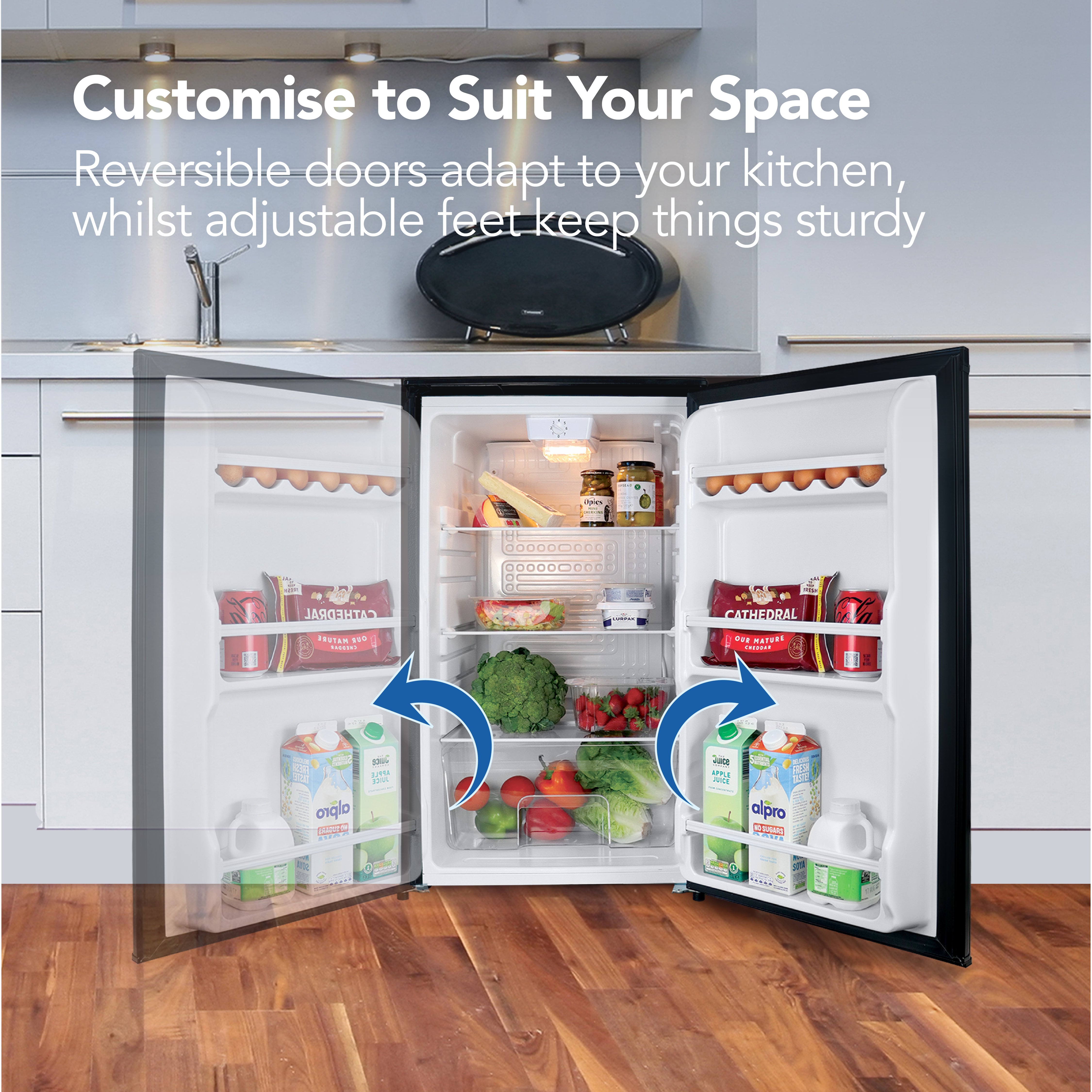 The Igenix Under Counter Larder Fridge (92L, Black) features a reversible door and adjustable feet for flexible placement. Shown in a modern kitchen with organized shelves full of food and drinks behind an open door.